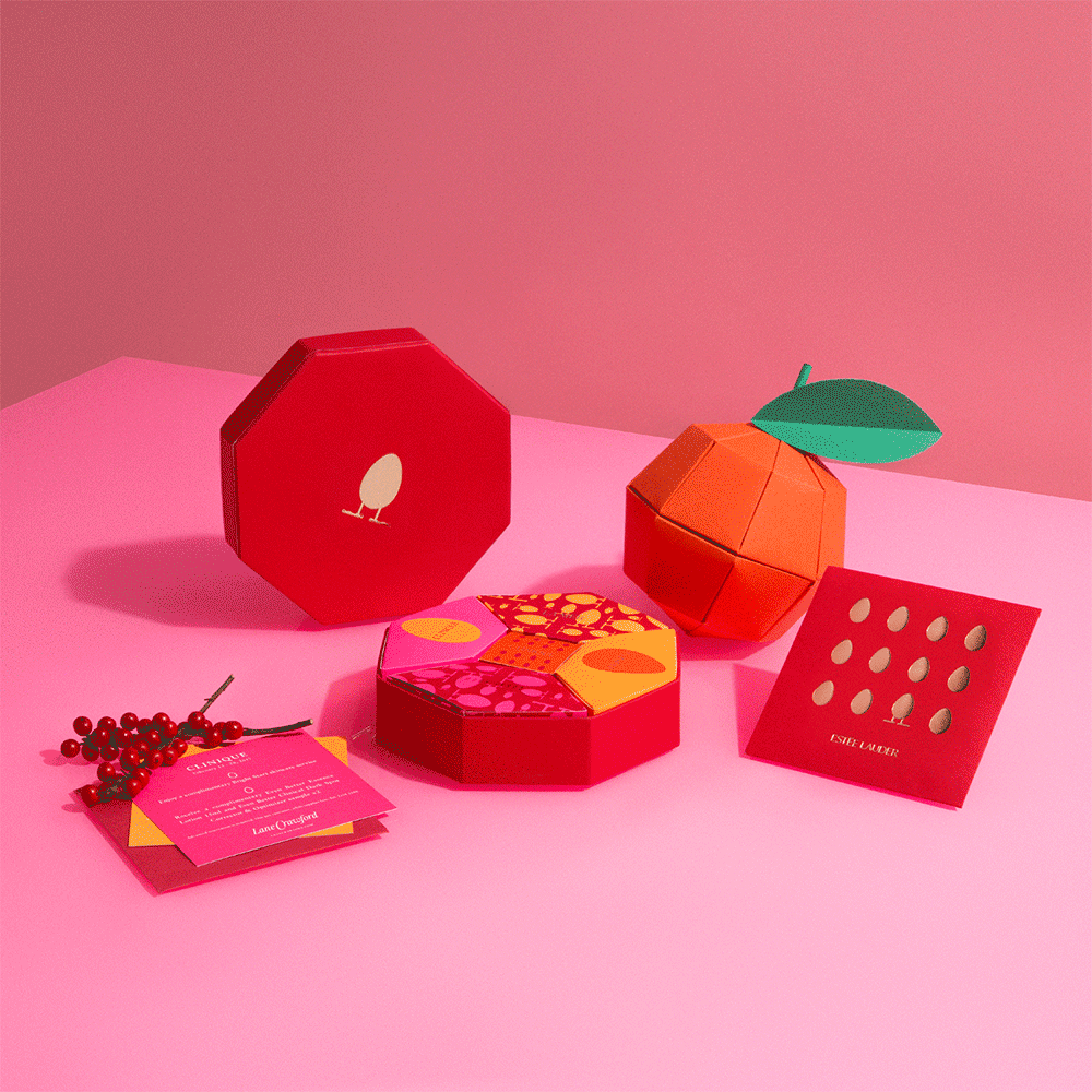 Colorful makeup and beauty products shaped like fruit on pink background, including an orange with a green leaf, a red purse with a bird illustration, a purple and yellow patterned makeup palette, a red card with hole patterns, and a pink card with t