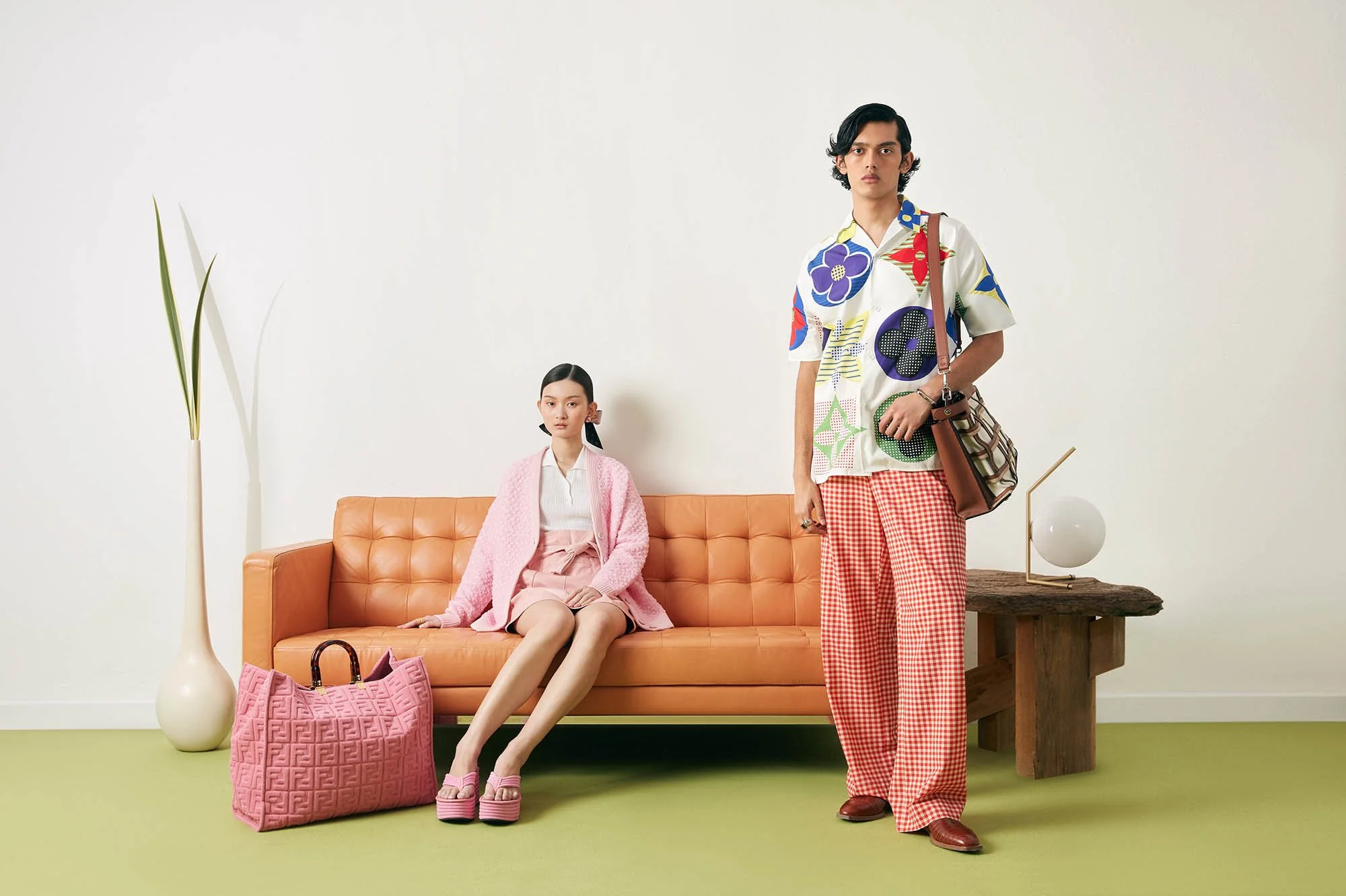 Two models in pastel-colored and patterned clothing pose in a minimalist room with a tan sofa, green carpet, and a wooden side table with a white lamp. One model sits on the sofa, the other stands beside it.