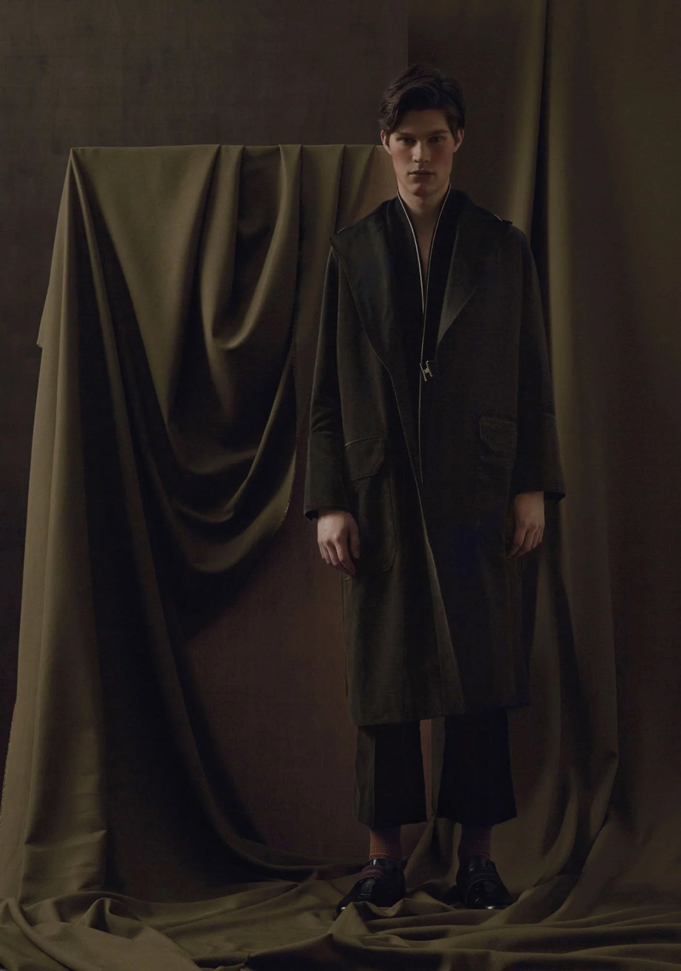 A young man stands in front of a dark background with draped fabric, wearing a long black coat, black pants, and black shoes.