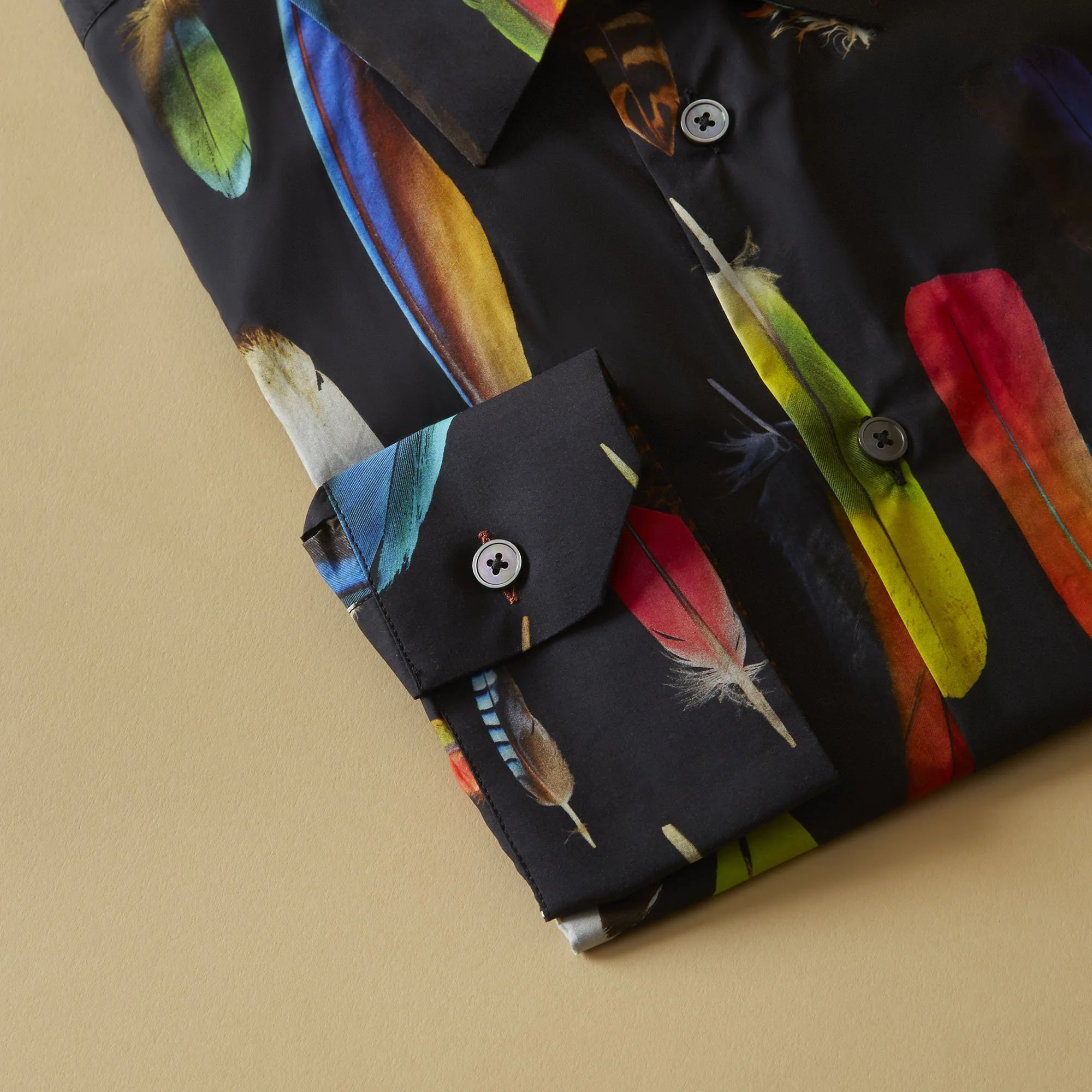 Close-up of a black button-up shirt with a colorful feather print design, folded neatly on a plain beige background.
