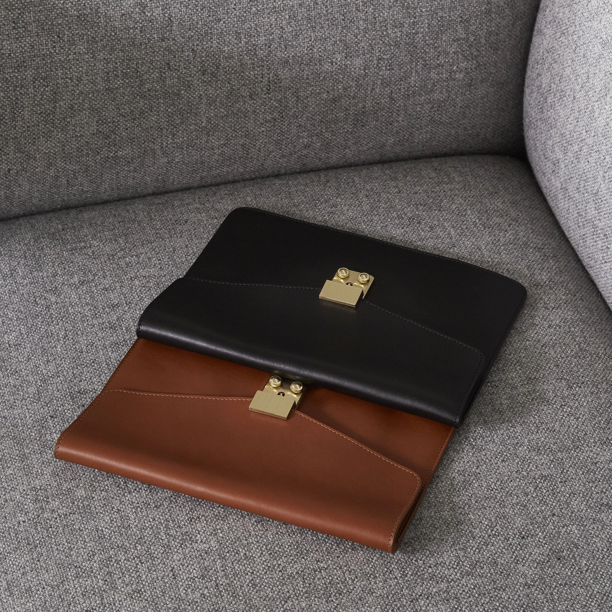 Two leather clutches, one black and one brown, with gold clasps, lying on a gray fabric sofa.