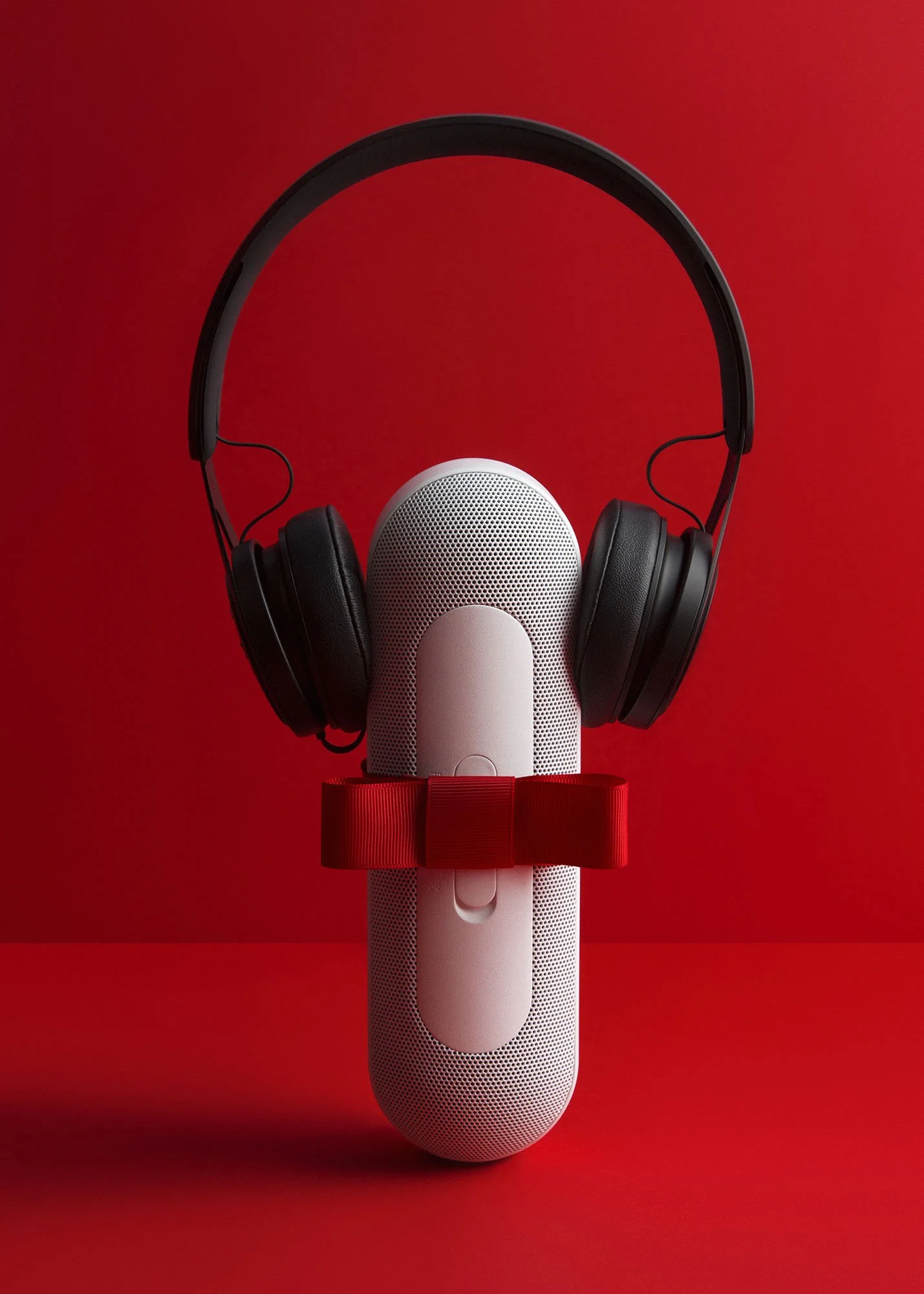 A white Google Nest Audio speaker with a red bow tie, topped with a pair of black over-ear headphones, against a red background.