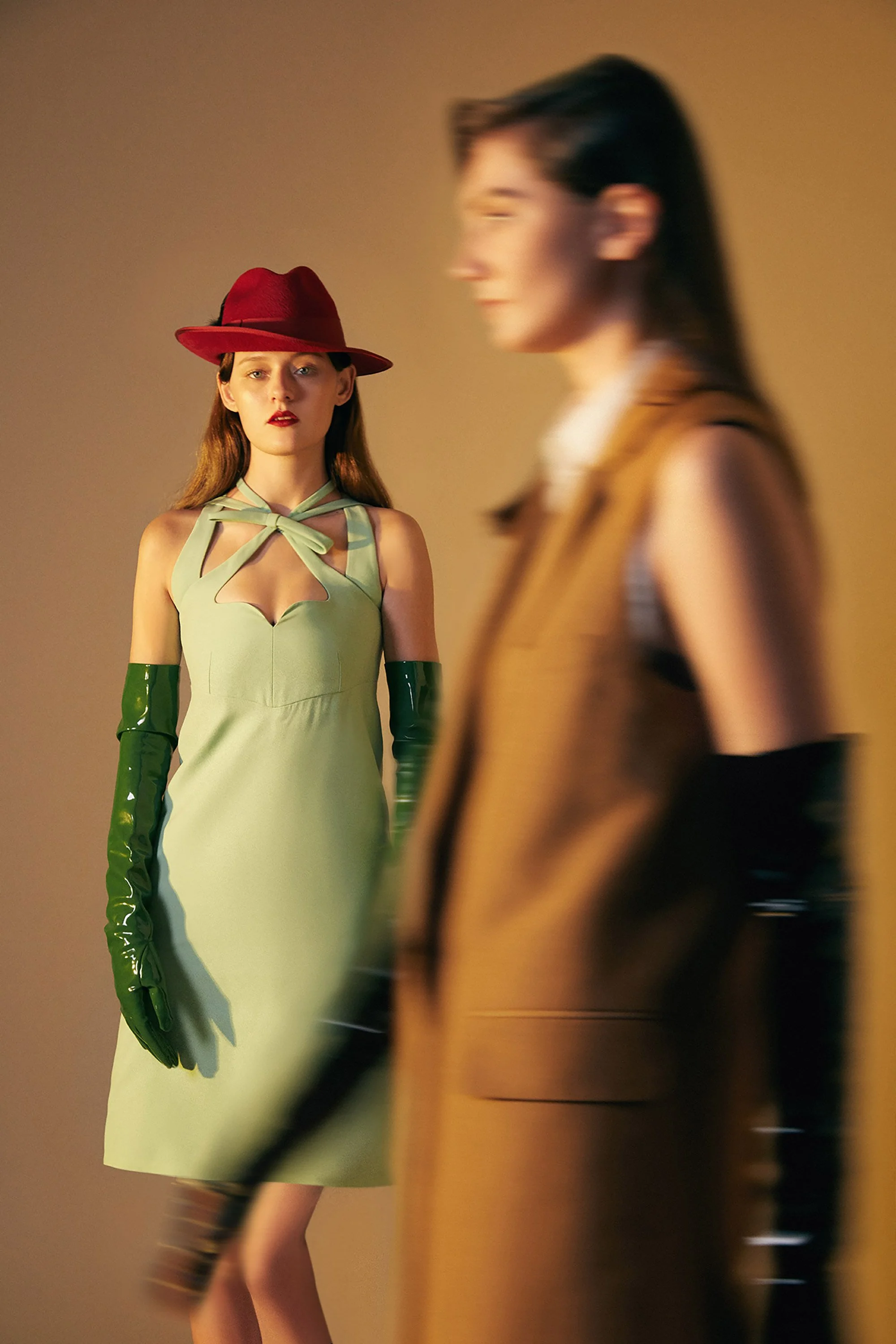 Two women in vintage-inspired fashionable outfits, one in the foreground wearing a tan coat and black gloves, and the other in the background wearing a green dress with a bow neckline, long green gloves, and a red hat, standing against a plain backgr