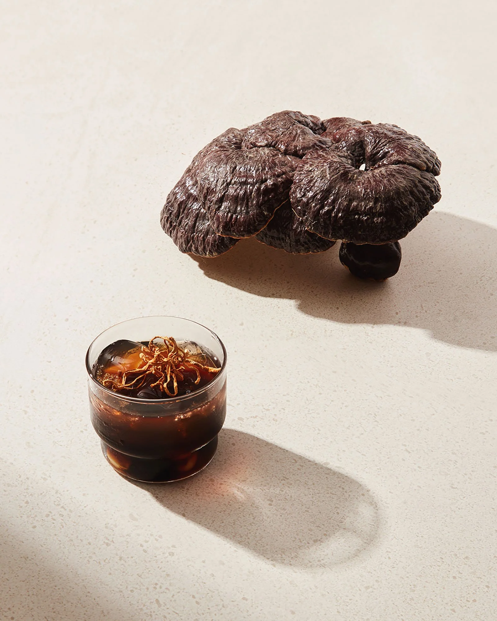 A dried black shiitake mushroom and a glass of dark soy sauce with sliced ginger on top, on a light-colored surface with shadows.