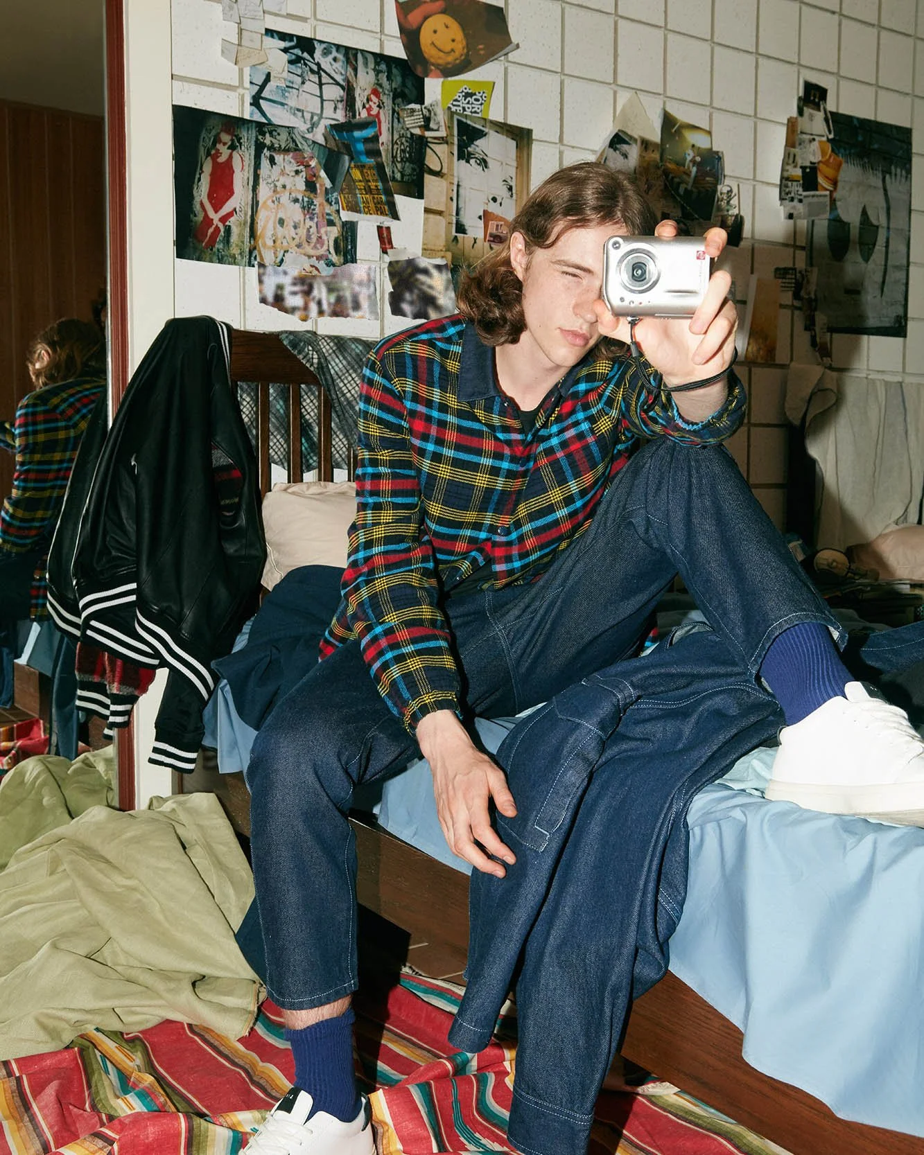 A person with wavy brown hair wearing a colorful plaid shirt, dark jeans, and white sneakers, taking a mirror selfie in a cluttered room. The room has a bed with scattered clothes, posters on the wall, and a striped rug.