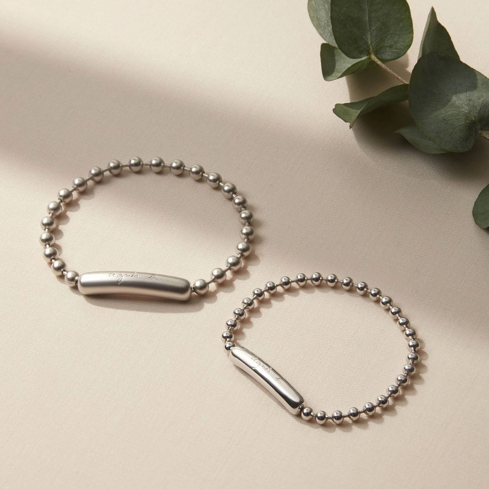 Two silver beaded bracelets with elongated oval clasps, placed on a light surface next to green leaves.