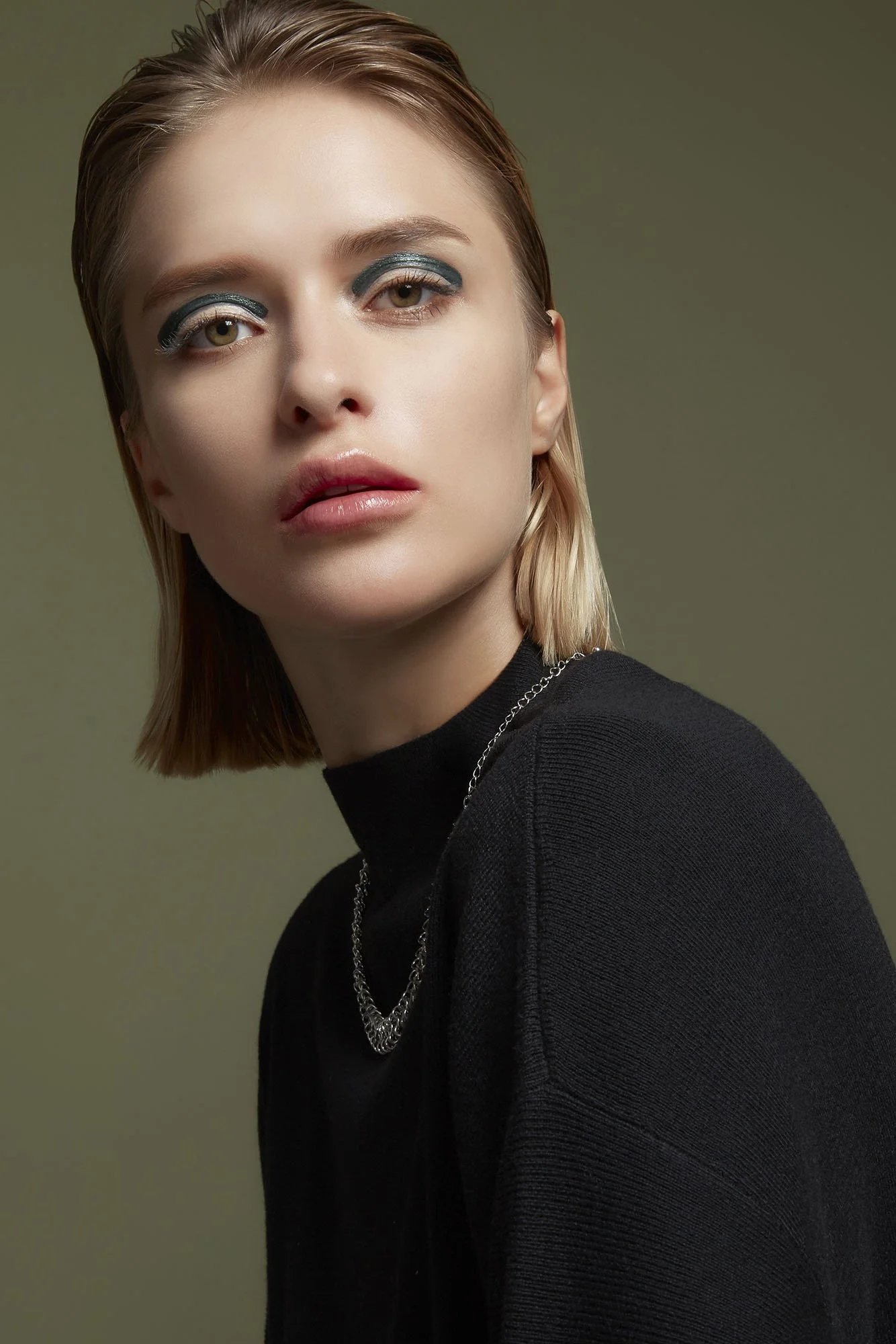 Close-up portrait of a young woman with short, blonde hair and bold eye makeup, wearing a black turtleneck and a silver chain necklace, against a muted green background.