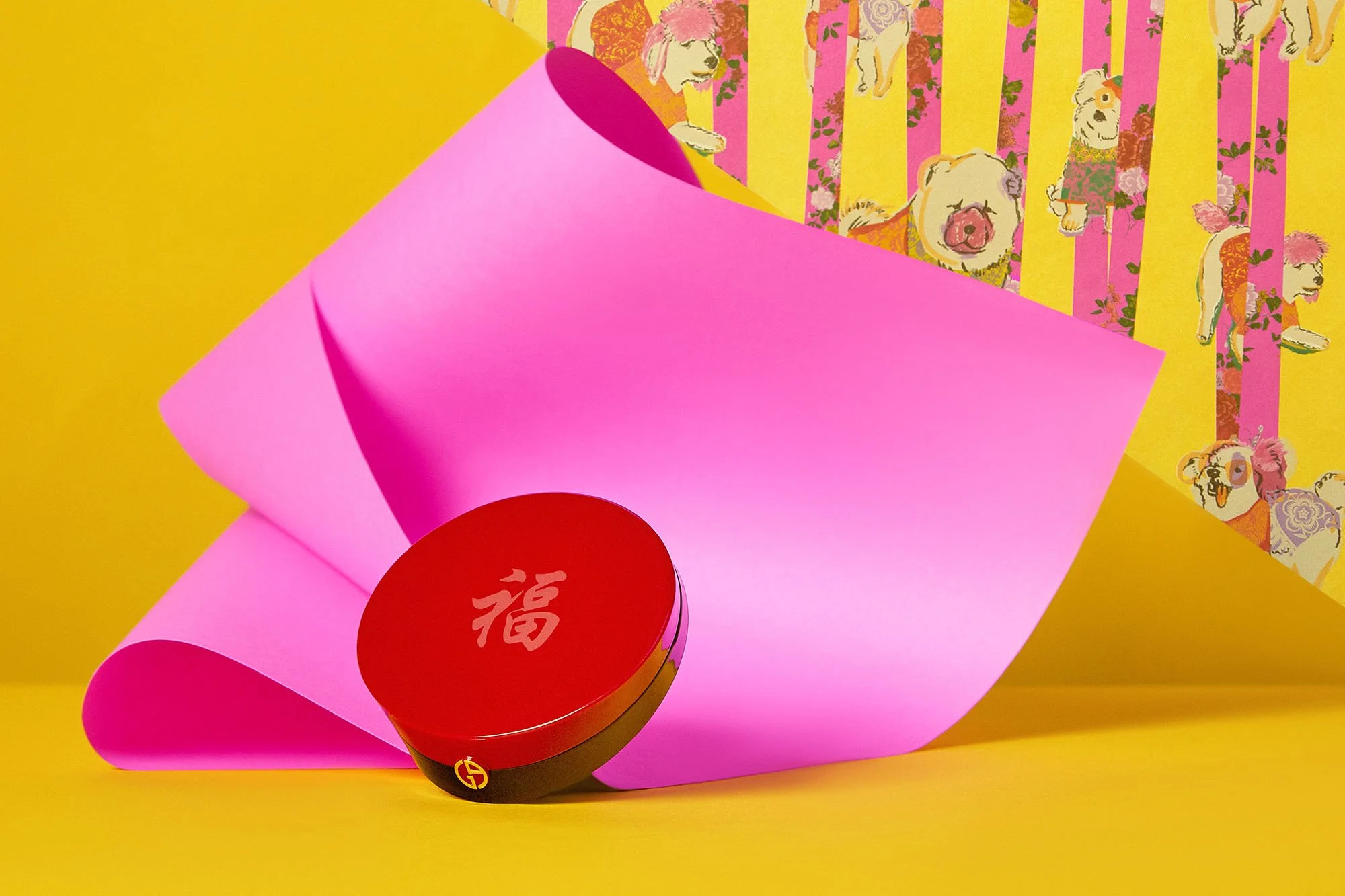 A pink and red circular box with a gold emblem sits on a yellow surface, with a pink paper sheet partially rolled and leaning behind it. In the background, there is wallpaper with illustrations of dogs in colorful outfits and floral patterns, with ve