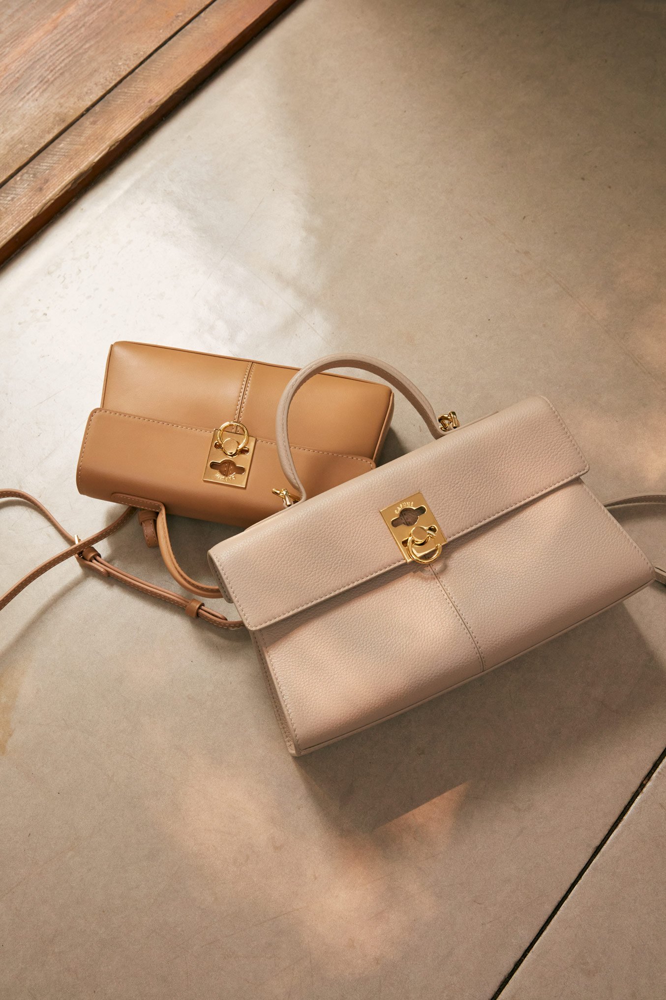 Three beige handbags with gold hardware placed on a gray tiled floor near a wooden surface.