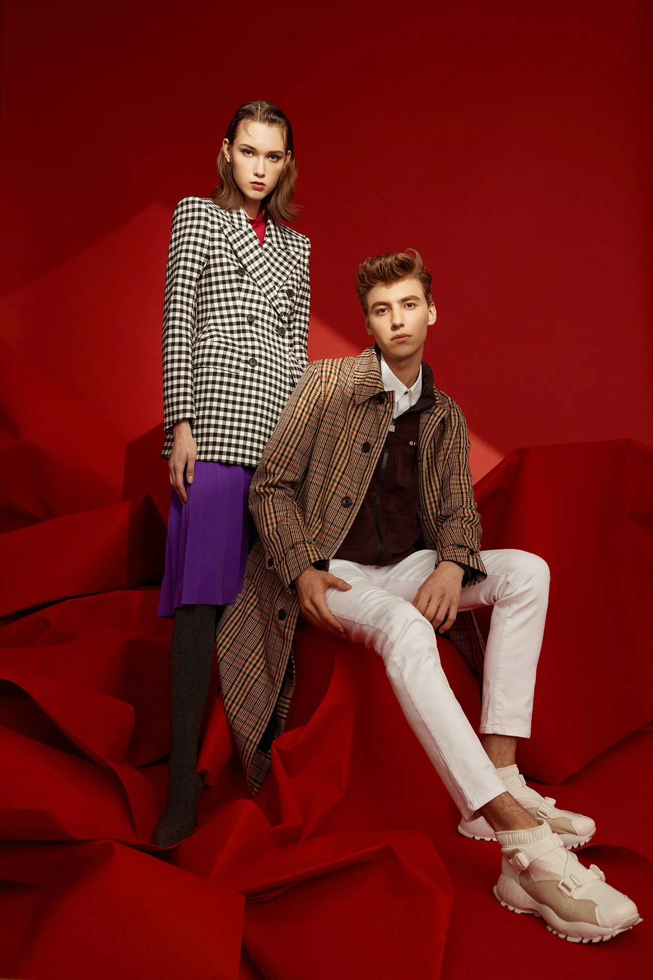 Two young models, a woman and a man, pose against a red background with red fabric. The woman wears a checkered blazer, purple skirt, and gray tights. The man wears a checked coat, white pants, and sneakers.