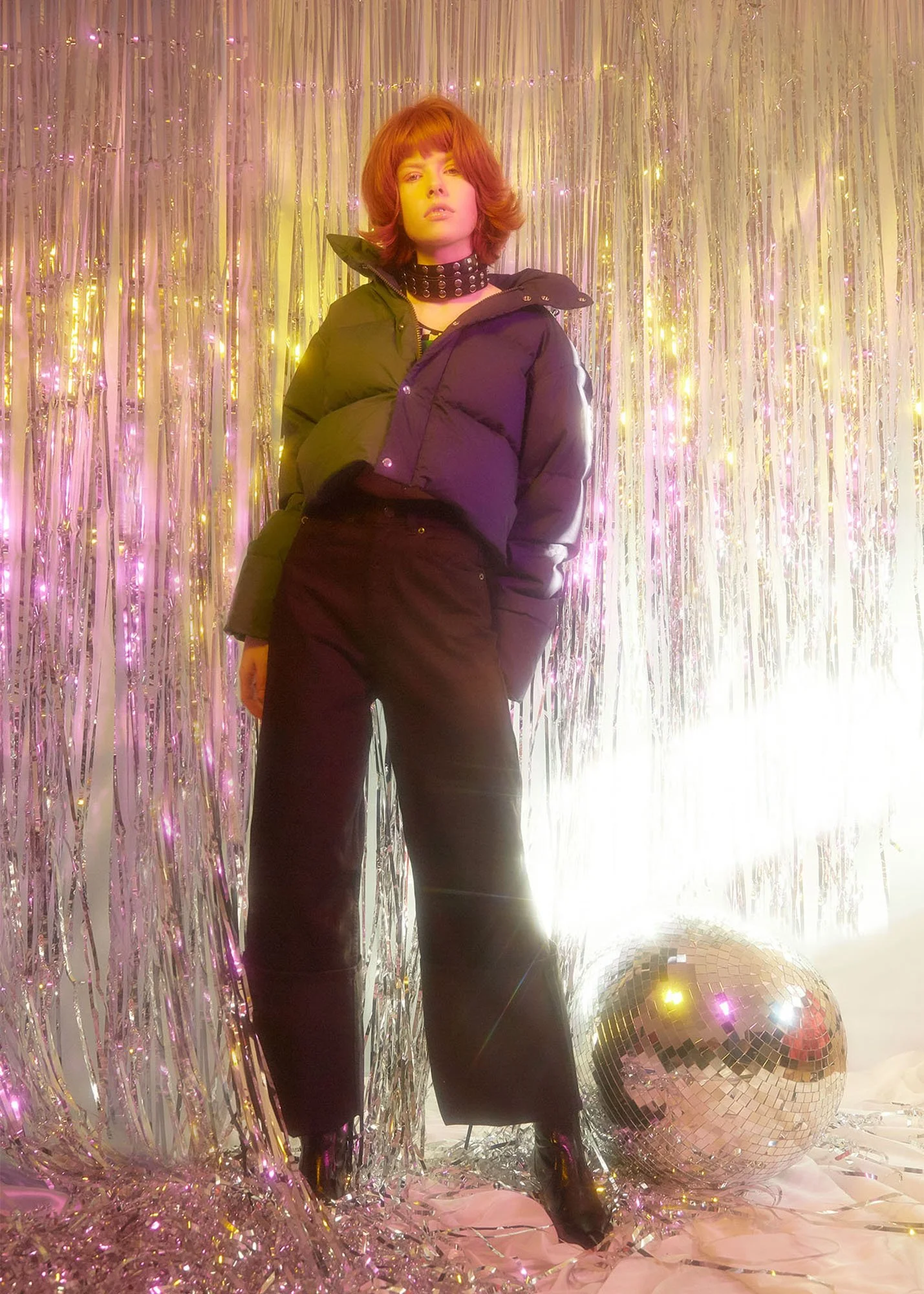 A woman with red hair wearing a black puffer jacket, black pants, and a studded choker, standing in front of a shimmering silver fringe backdrop with pink and yellow lighting, next to a disco ball on the floor.