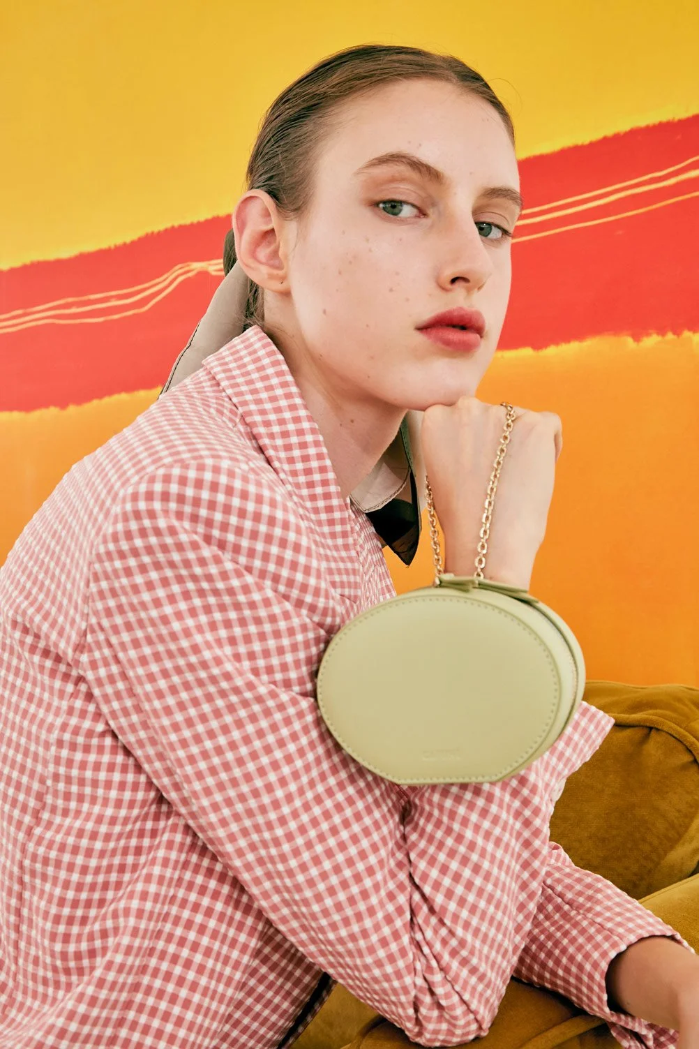 Young woman with light skin and red hair, wearing a pink and white checkered blazer, sits on a tan couch with a yellow background featuring an abstract red and orange design, holding a small oval-shaped yellow handbag with a chain strap.
