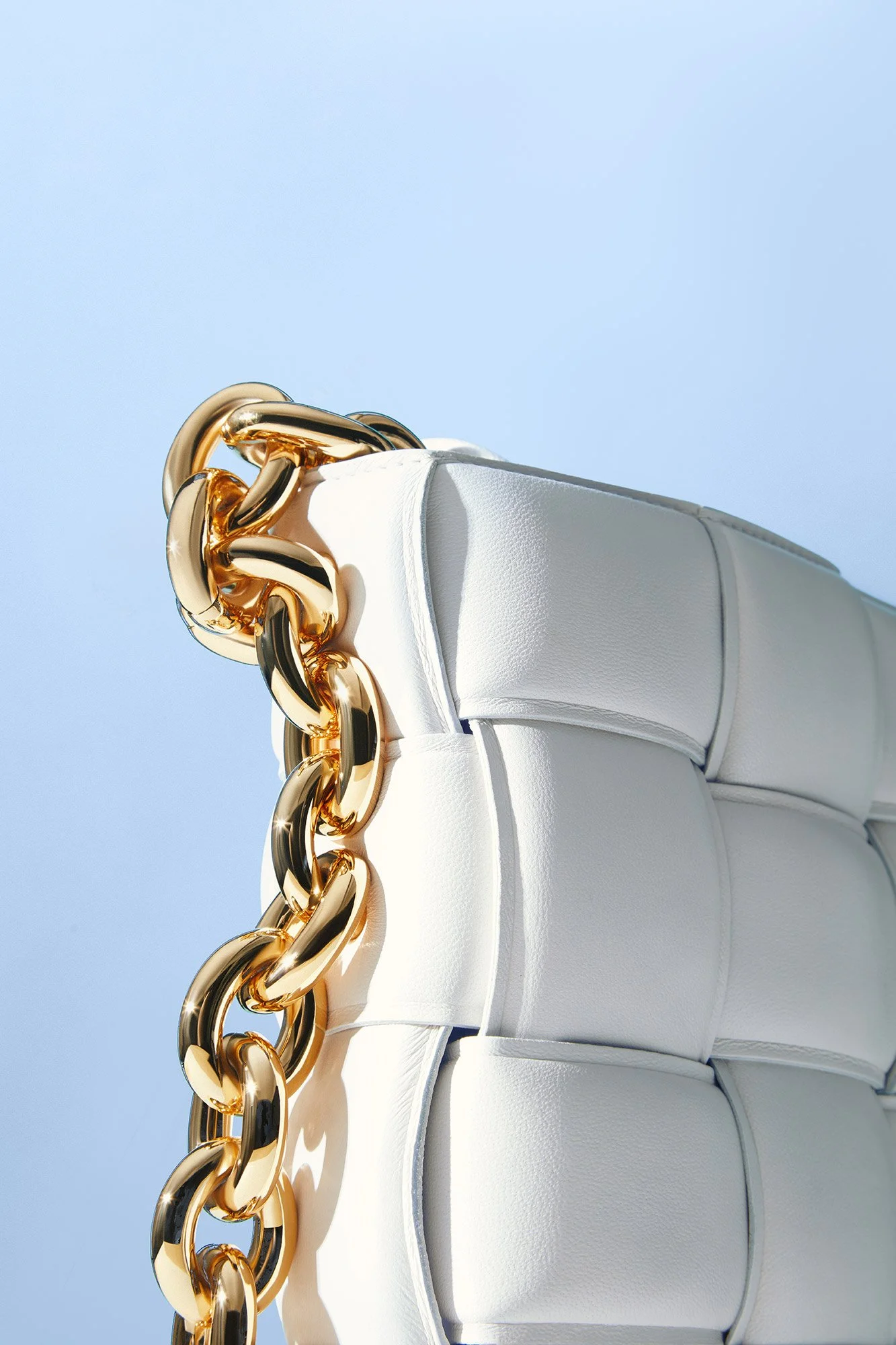 Close-up of a white handbag with a gold chain strap against a clear blue sky.