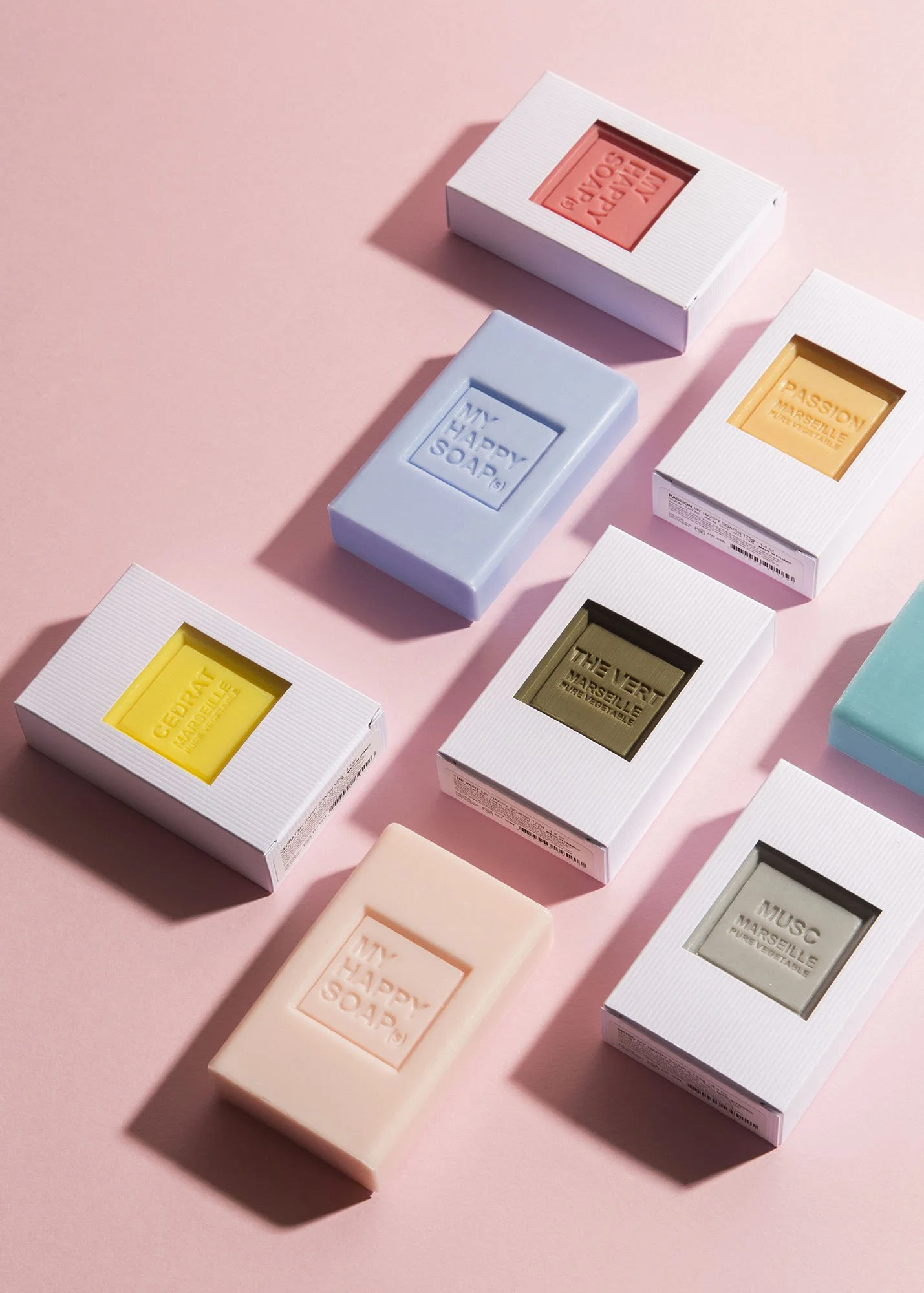 Colorful soap bars in pastel shades with white packaging and embossed text, arranged on a pink background.