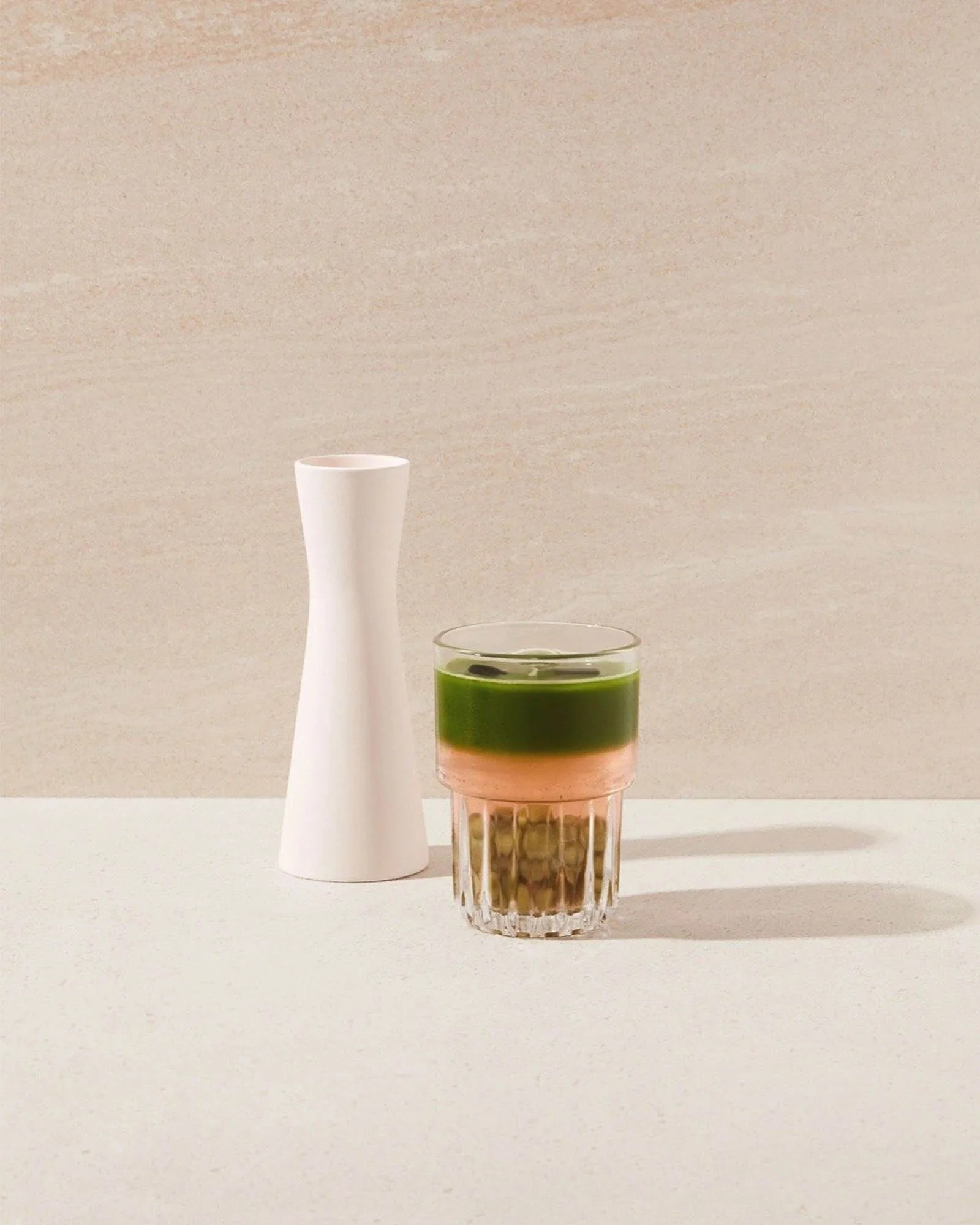 A white ceramic vase and a glass containing a layered beverage with green, pink, and brown layers on a white surface against a beige background.