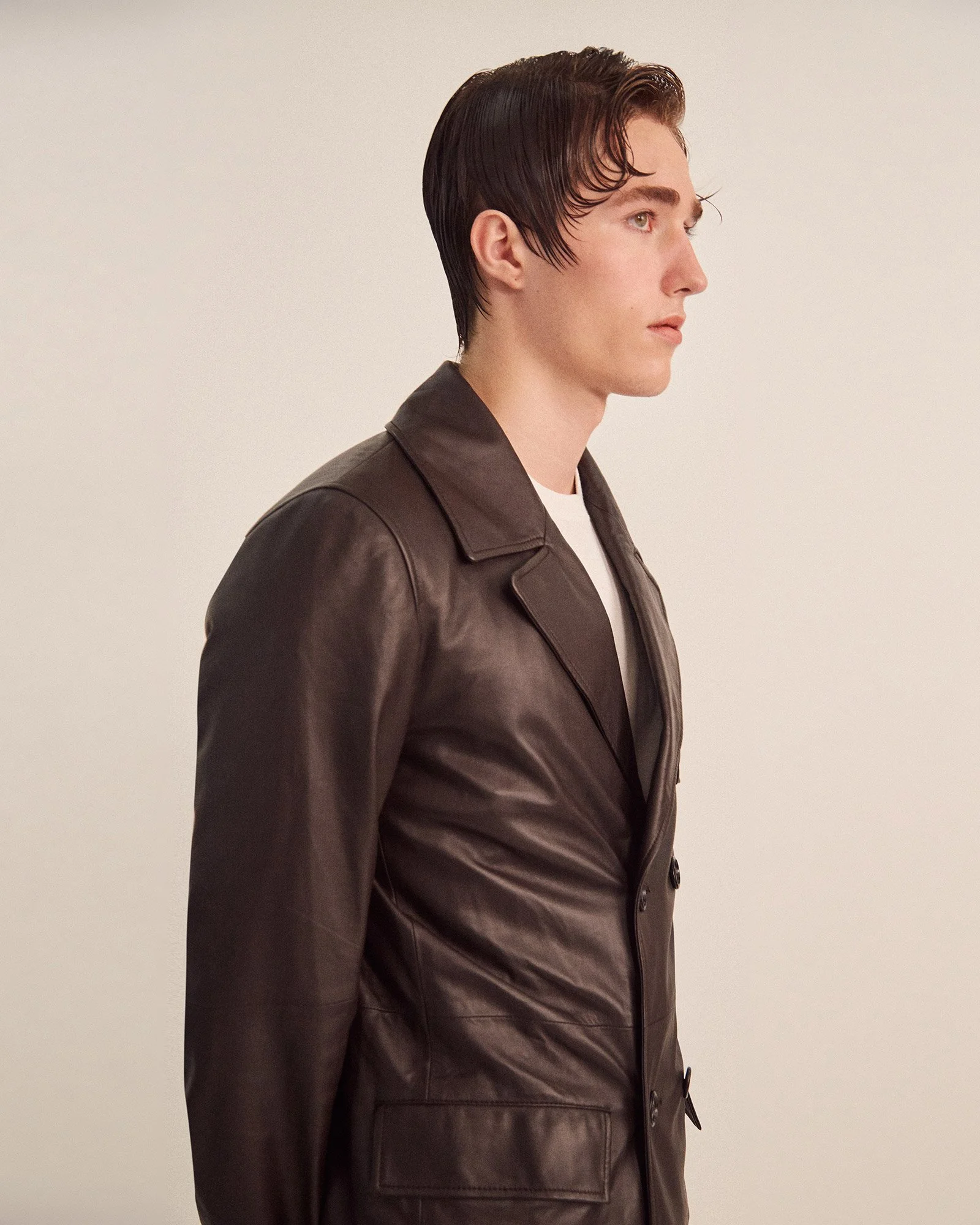 Side profile of a young man with wet, dark hair, wearing a dark brown leather jacket over a white shirt, standing against a plain, light-colored background.