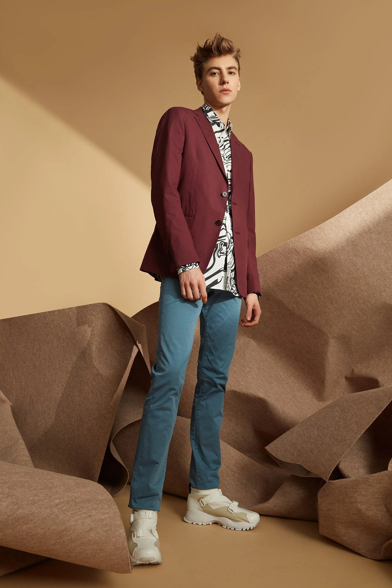 A young man standing in a studio with a beige background and large brown paper-like props. He is wearing a maroon blazer over a black and white patterned shirt, light blue pants, and off-white sneakers.