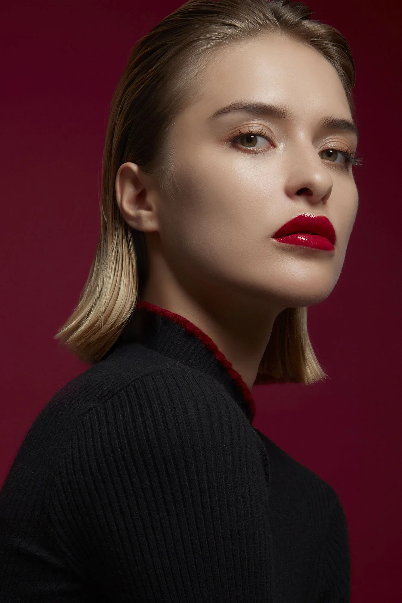 Close-up portrait of a woman with blonde hair, wearing red lipstick, and a high-neck black sweater, against a red background.