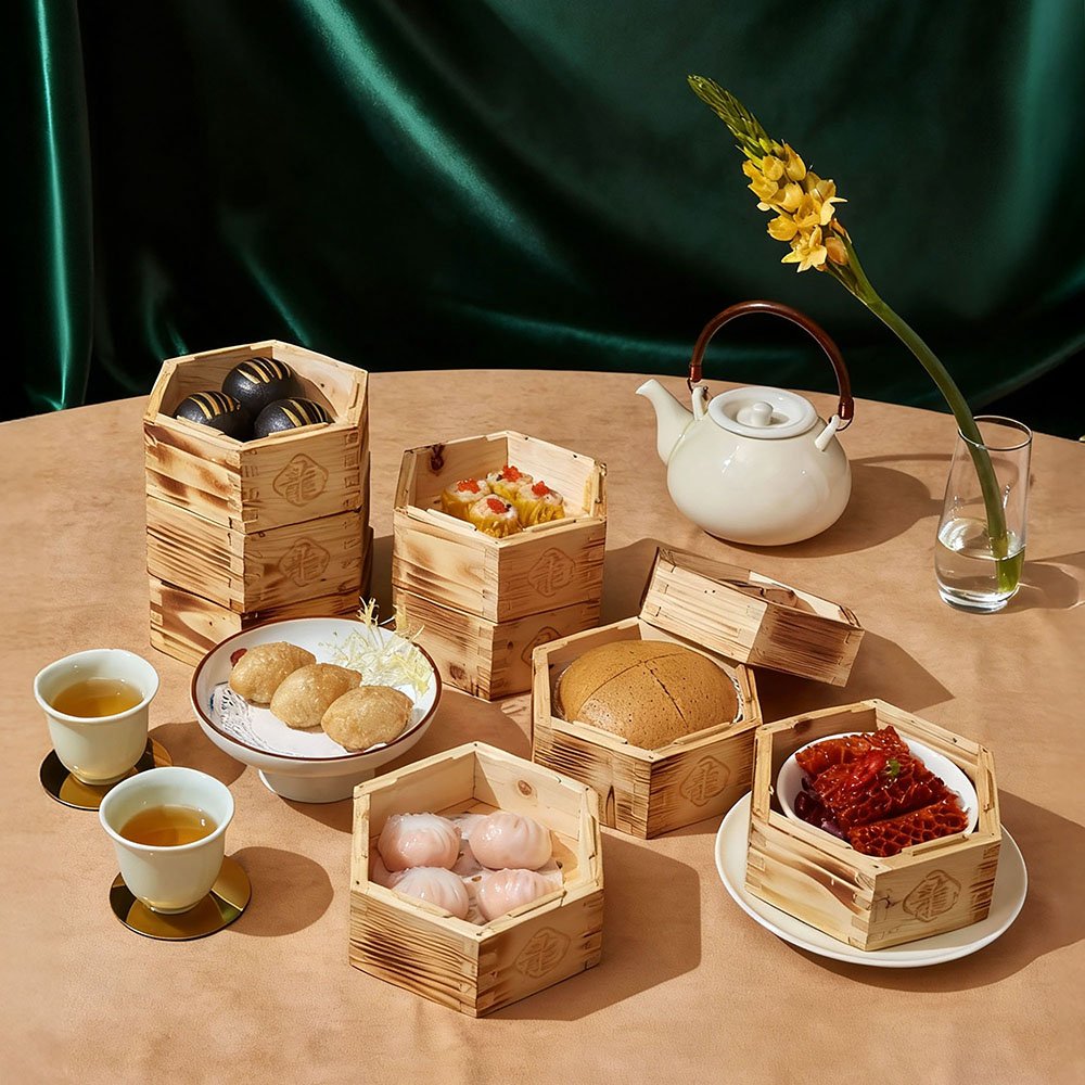 Tea service with assorted dim sum, desserts, and tea cups on a wooden table with a green backdrop and a tall flower in a glass vase.
