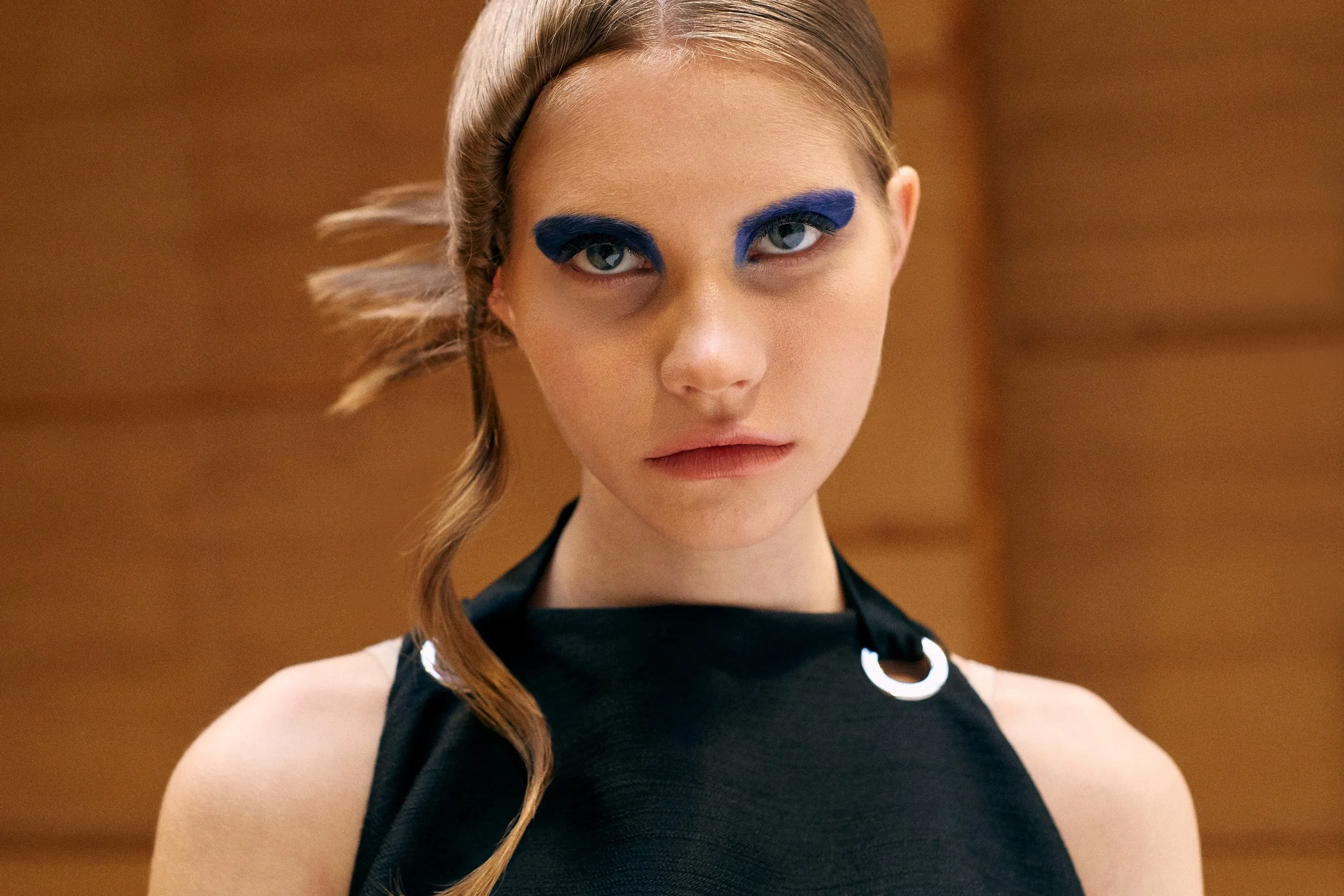A young woman with blonde hair styled in a side braid, wearing bold blue eye makeup and a black sleeveless top with circular metallic details, looking directly at the camera against a wooden background.