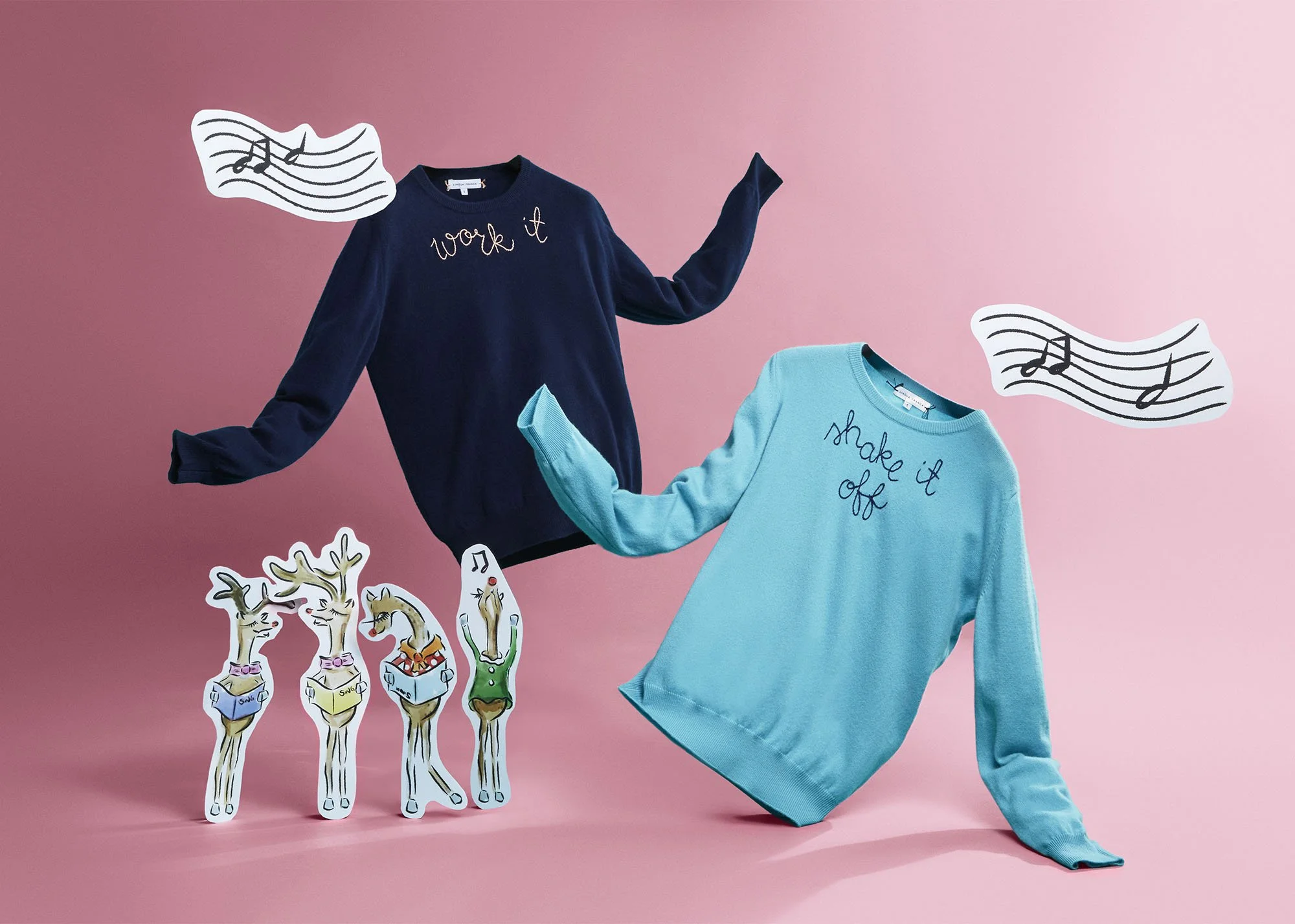 Two sweaters, one navy with the words 'work it' and one light blue with the words 'shake it off,' are displayed against a pink background with musical notes and three reindeer stickers.