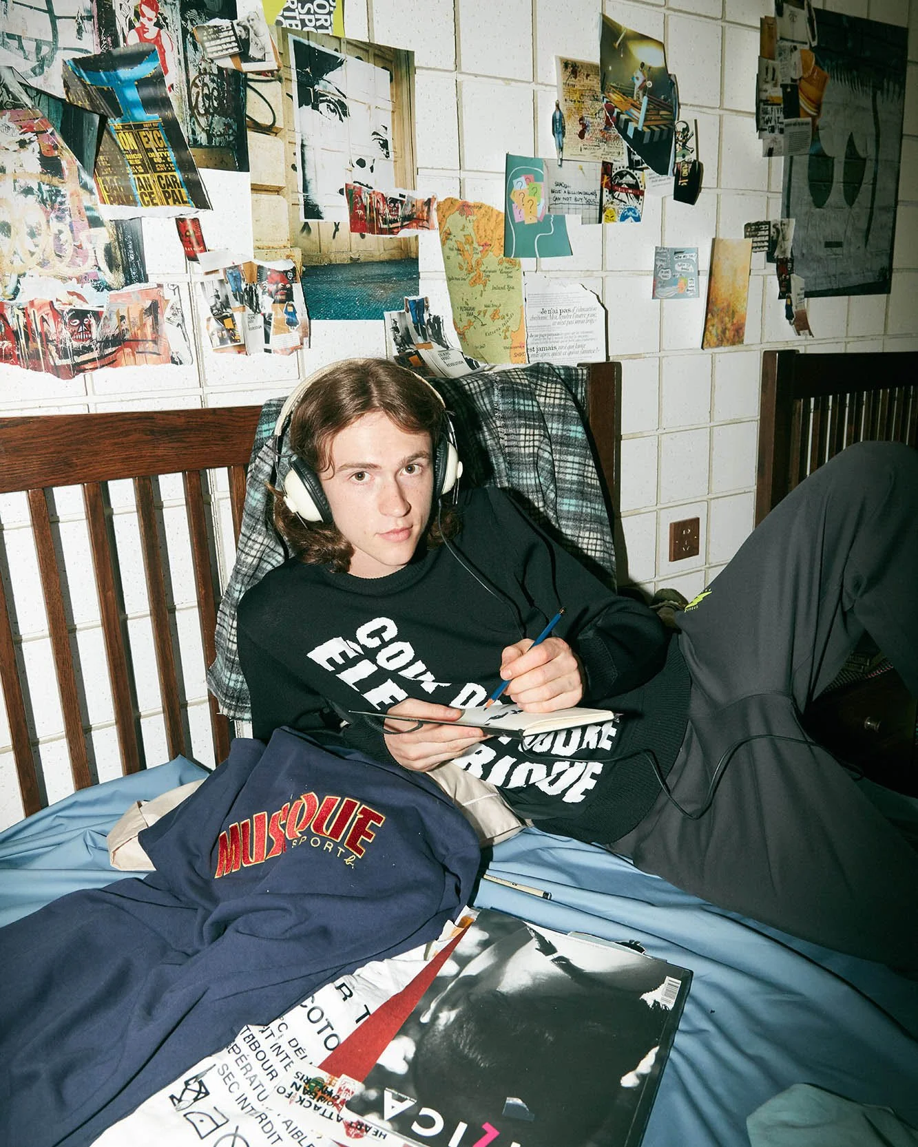 A young man with shoulder-length brown hair wearing headphones, a black sweatshirt, and dark pants, lying on a bed in a room with a cluttered wall of posters, magazine clippings, and artwork behind him. He is holding a notebook and pen, surrounded by