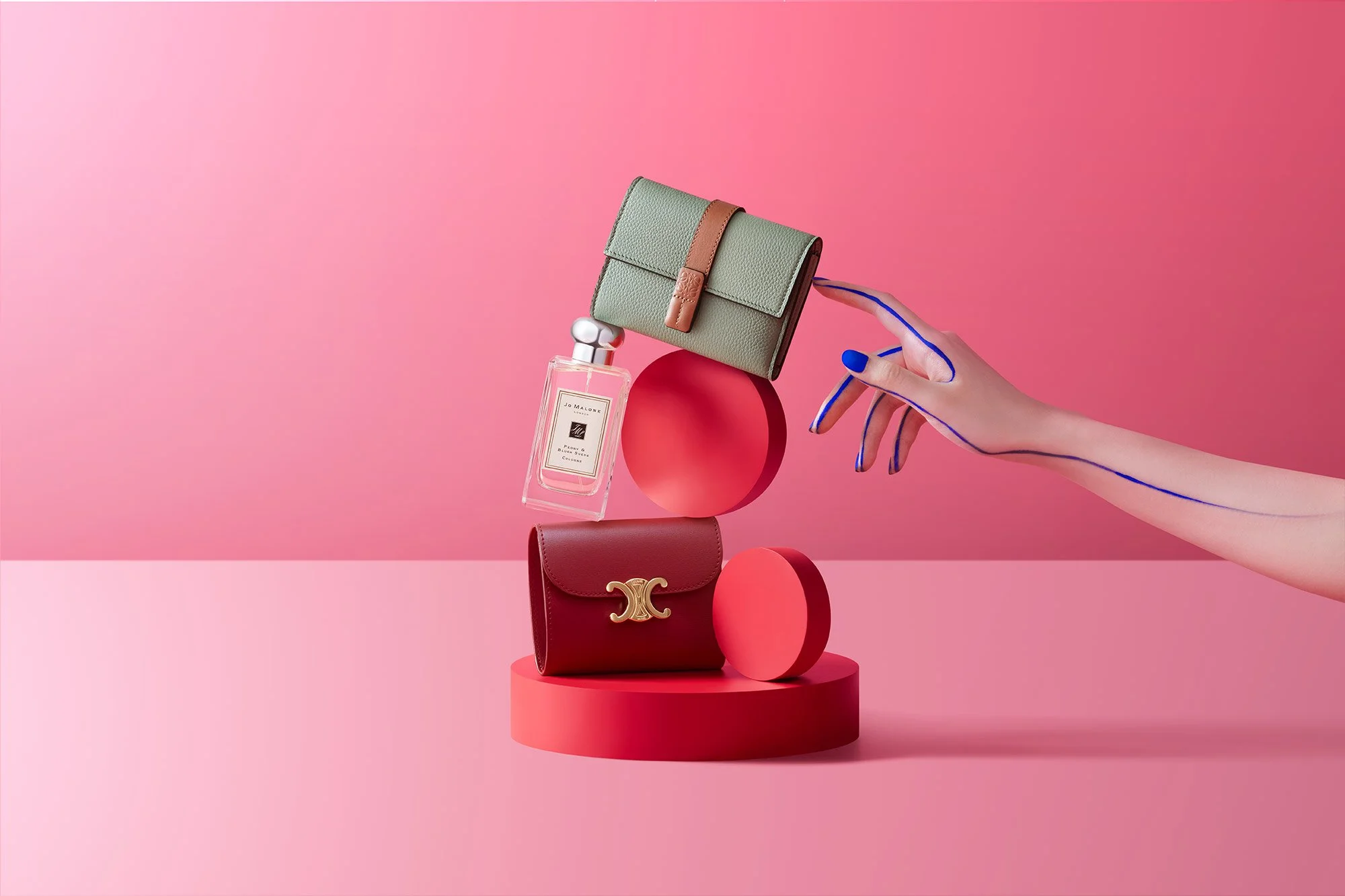 Display of luxury accessories and perfume bottle on pink geometric background, with a hand pointing at the items.