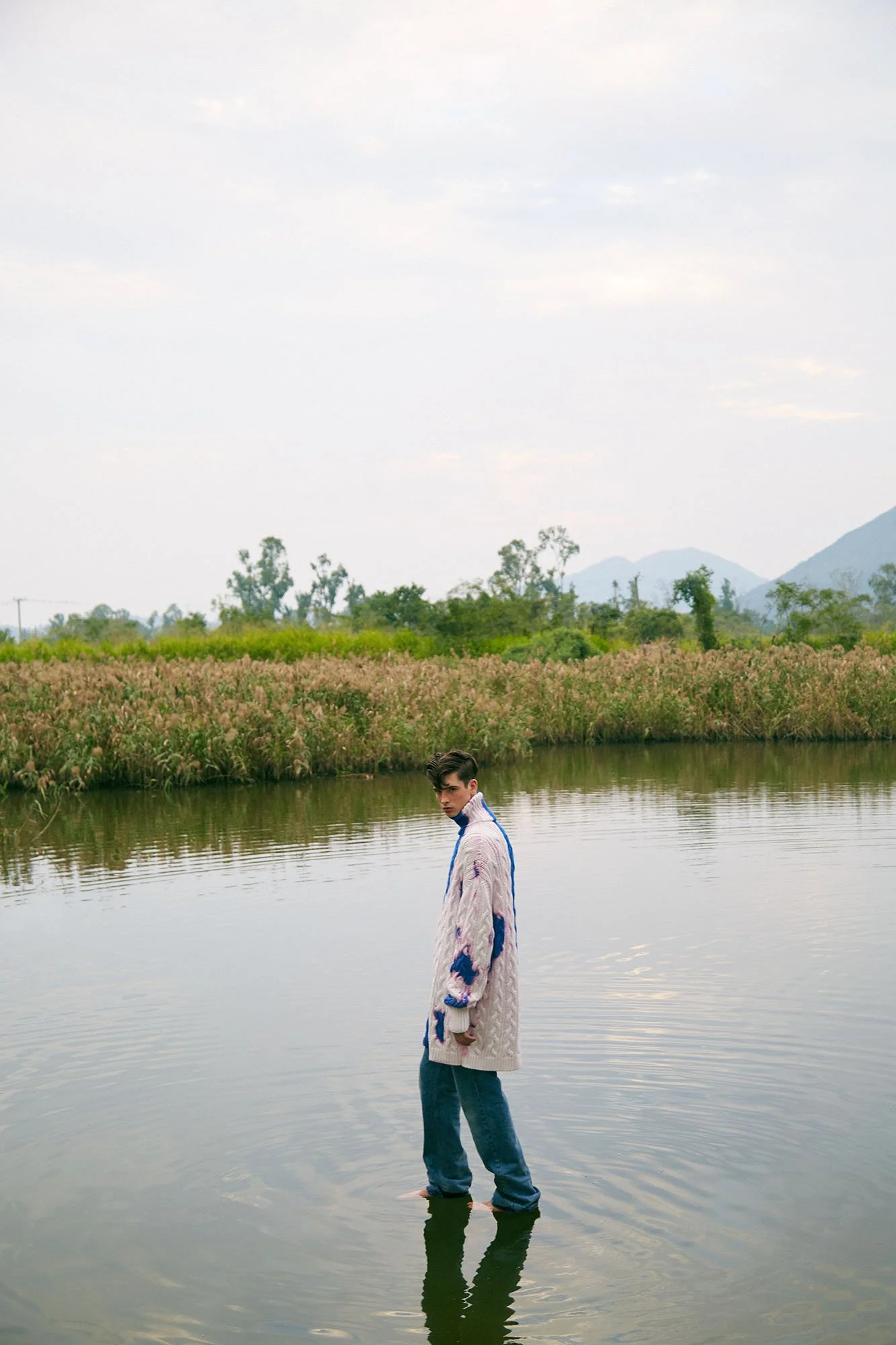 A young man standing in a calm body of water, wearing a long, cozy sweater and jeans, with a natural landscape, trees, and mountains in the background under a cloudy sky.