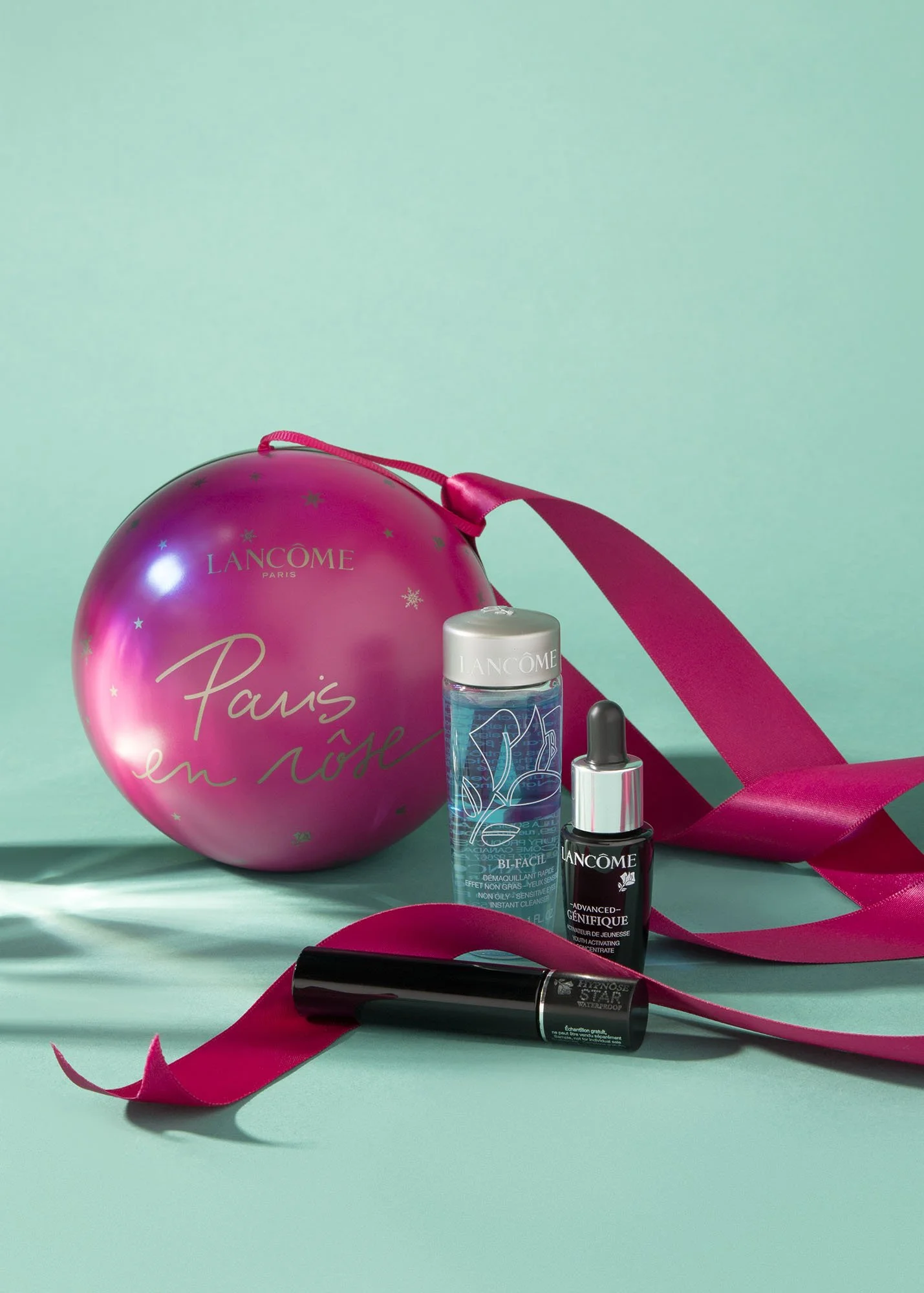 A pink Christmas ornament with gold and silver details, a pink ribbon, and three skincare products from Lancôme in front of a pastel background.