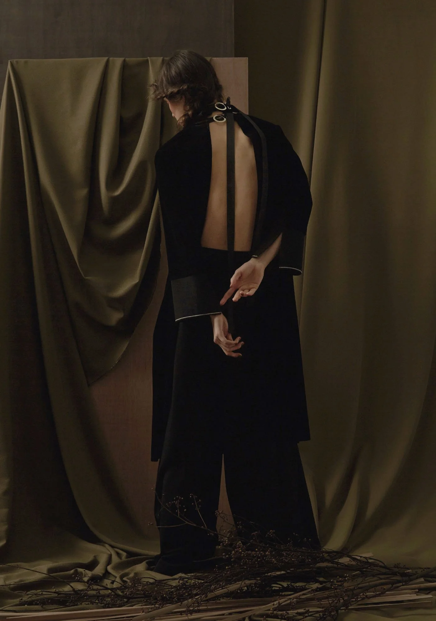 A woman dressed in a backless black outfit stands with her back to the camera, in front of dark curtains and draped fabric, with dried branches on the floor.