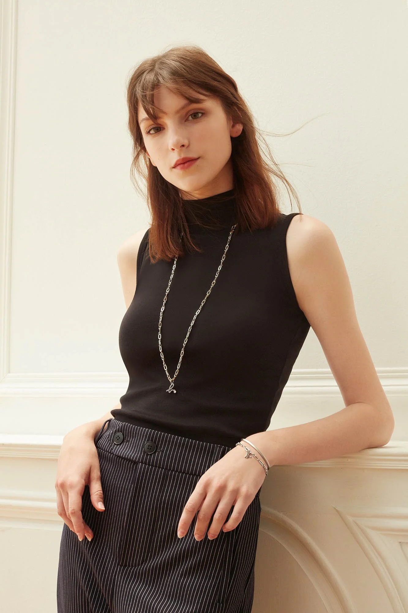Young woman with shoulder-length brown hair wearing a sleeveless black turtleneck top, pinstripe pants, and jewelry including a necklace and bracelets, leaning against a white wall with decorative molding.