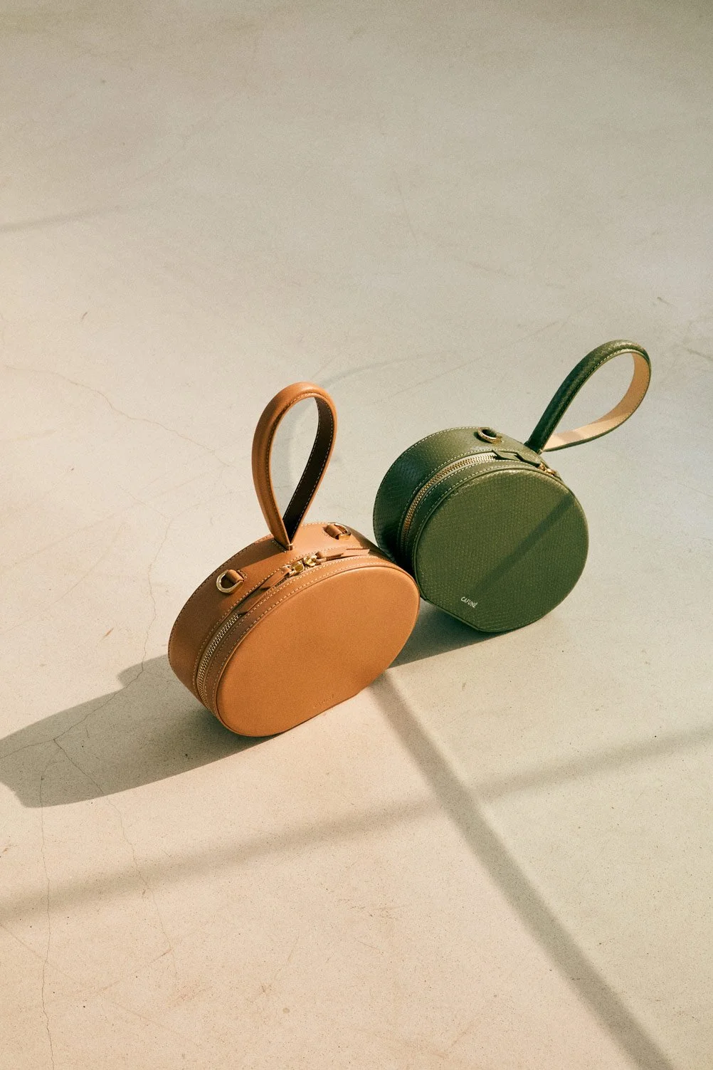 Two round handbags, one tan and one green, with handles and zippers, placed on a plain surface with shadows.