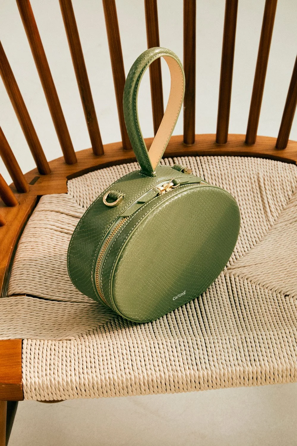 A green, textured, round handbag with gold zippers and a handle, placed on a woven beige cushion on a wooden chair with vertical slats.