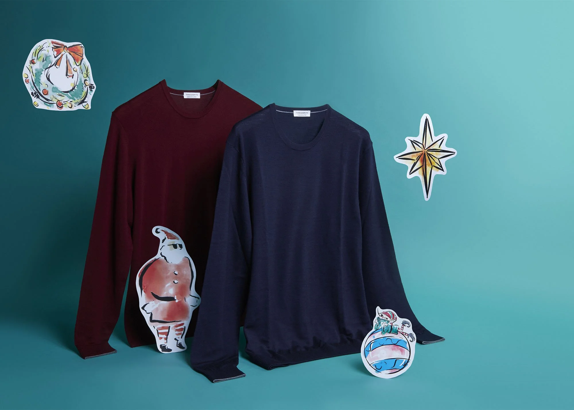 Two long-sleeve shirts, one red and one navy blue, are hanging against a teal background, with colorful holiday-themed paper cutouts of a Christmas ornament, a star, Santa Claus, and a wrapped gift surrounding them.