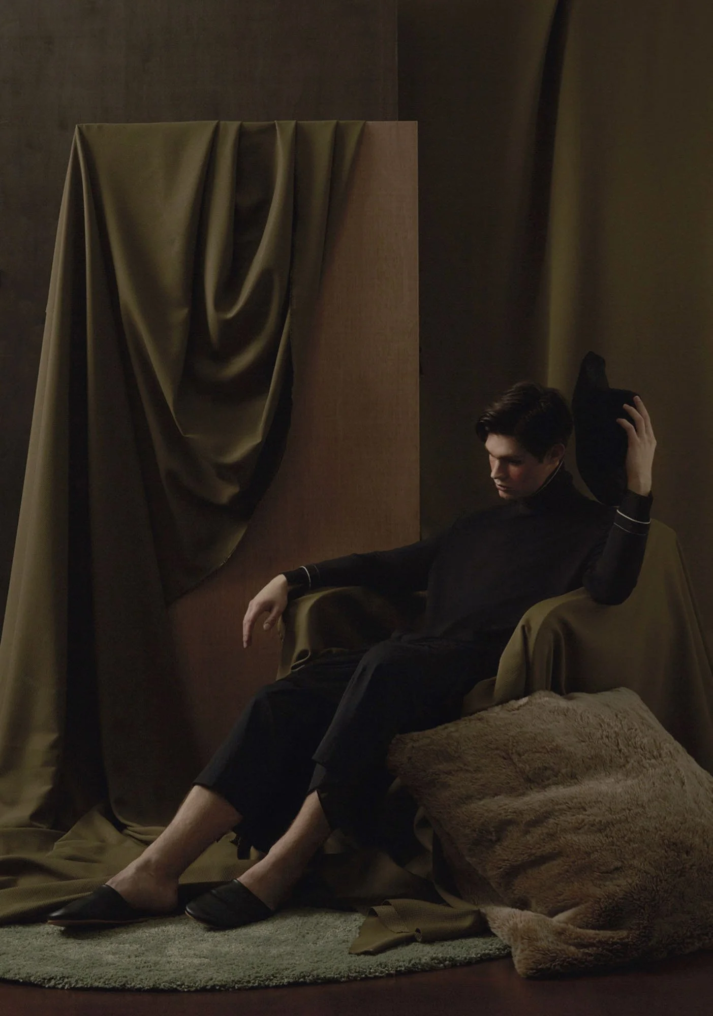 A young man seated in a brown armchair, wearing black clothing, with one arm resting on the chair's armrest and the other holding a black hat, surrounded by dark brown and green drapes and cushions in a dimly lit room.
