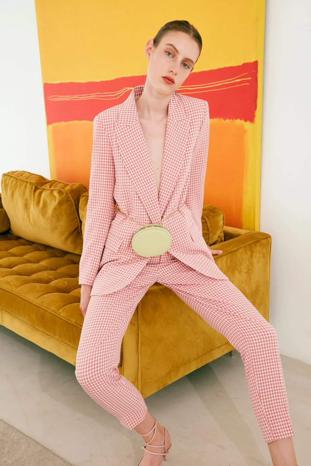 A woman wearing a pink and white checkered suit with a deep V neckline, sitting on a mustard yellow velvet sofa. She has short brown hair and is accessorized with a small round purse hanging on her waist. Behind her, there is an abstract yellow and r
