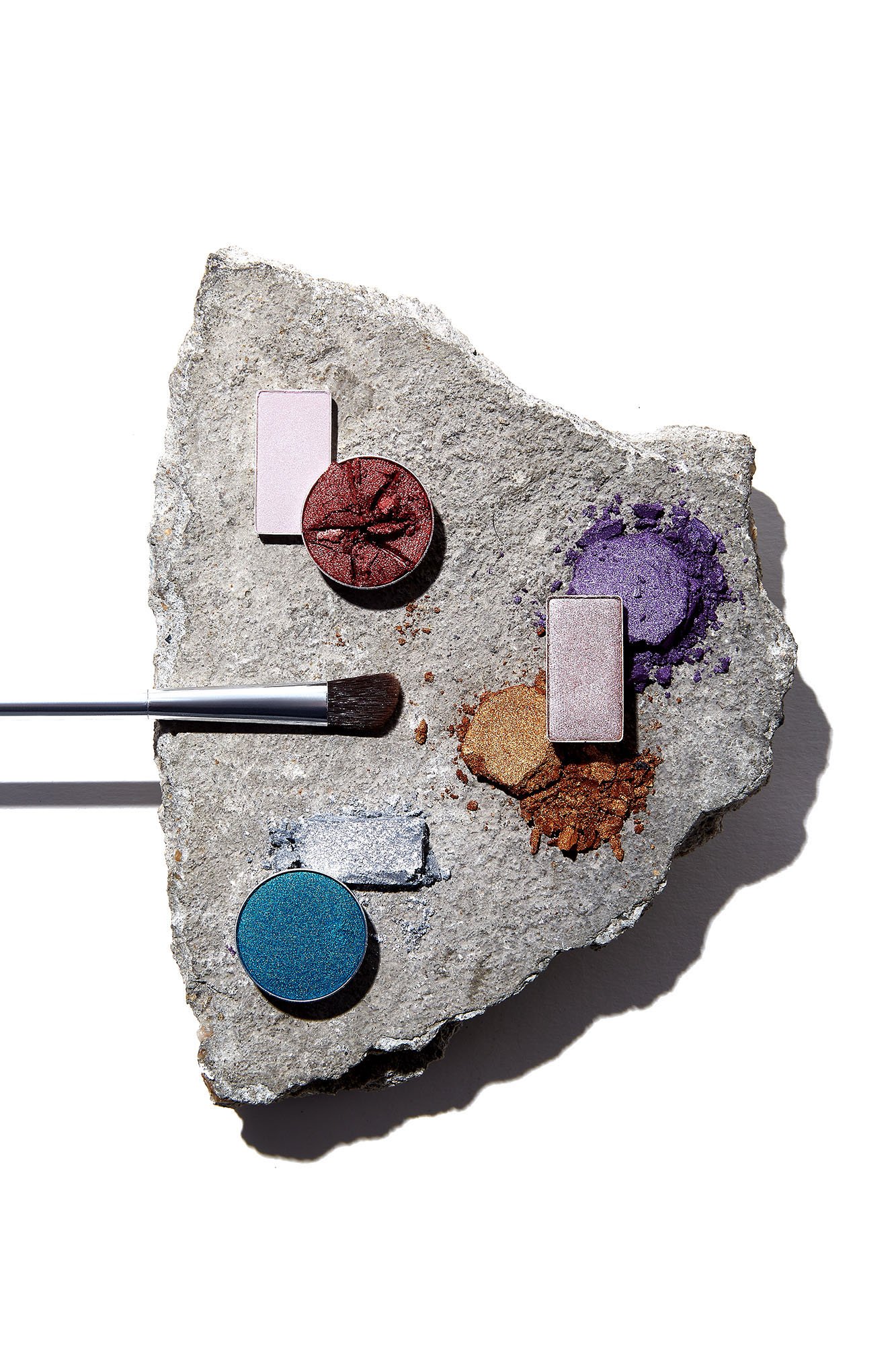 Makeup eyeshadow palette with brushes on a rough stone surface featuring colorful eyeshadow shades in pink, red, purple, blue, and gold.