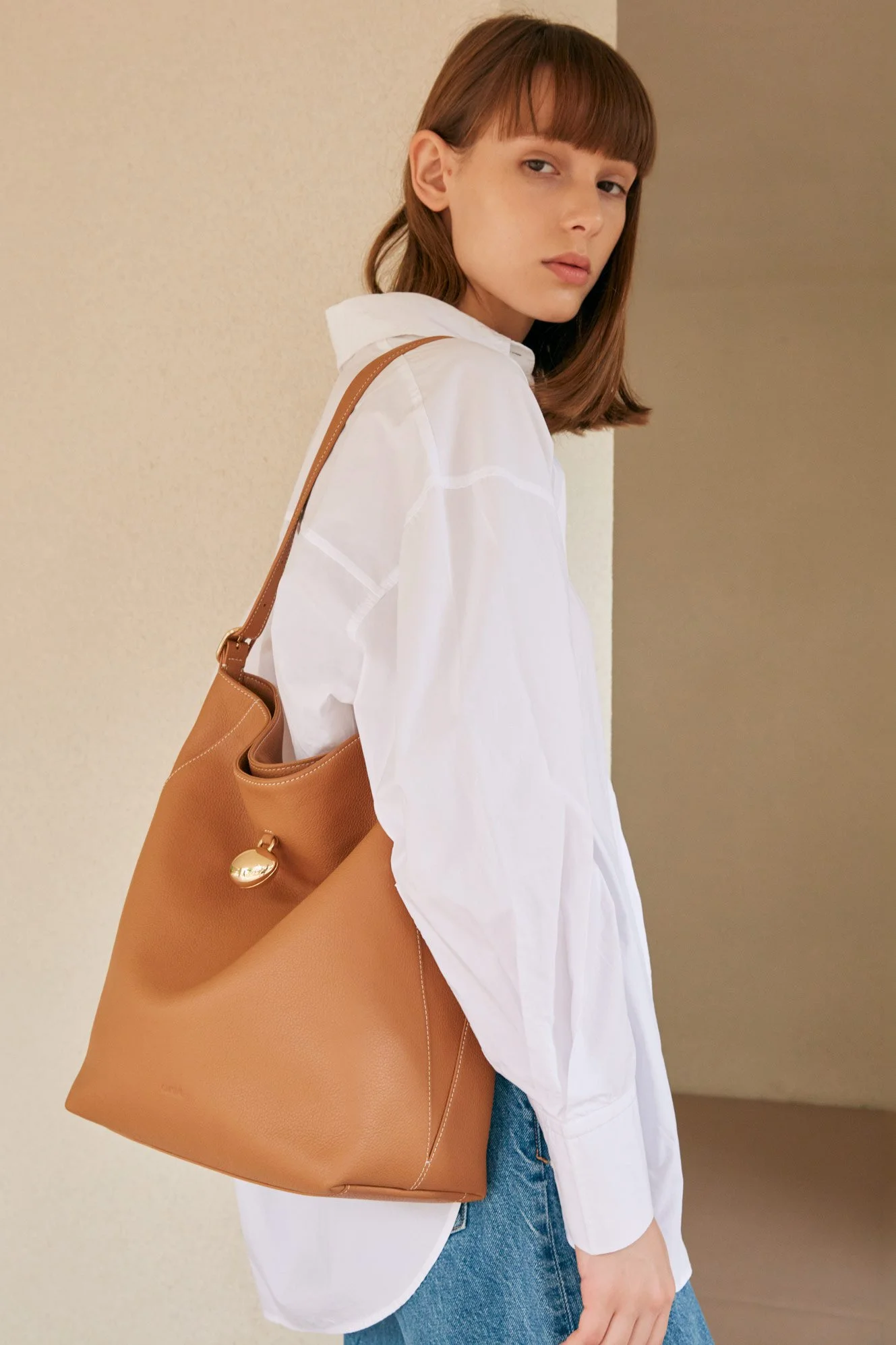 A young woman with brown hair and bangs, wearing a white oversized shirt and blue jeans, is standing against a neutral background. She has a large tan leather shoulder bag with gold hardware.