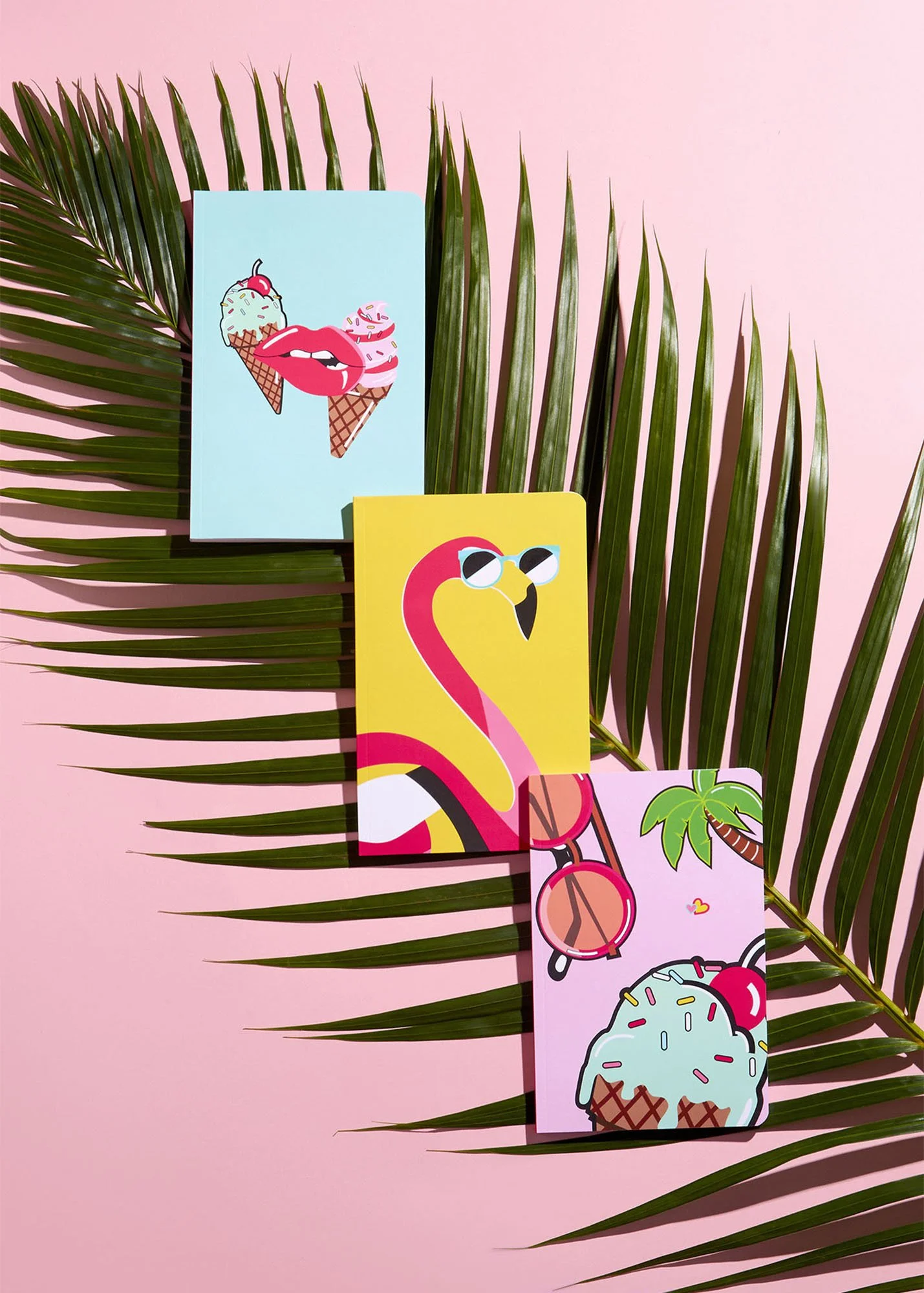 Colorful tropical-themed greeting cards with ice cream, flamingo, palm tree, and sunglasses designs, arranged on a large palm leaf against a pink background.