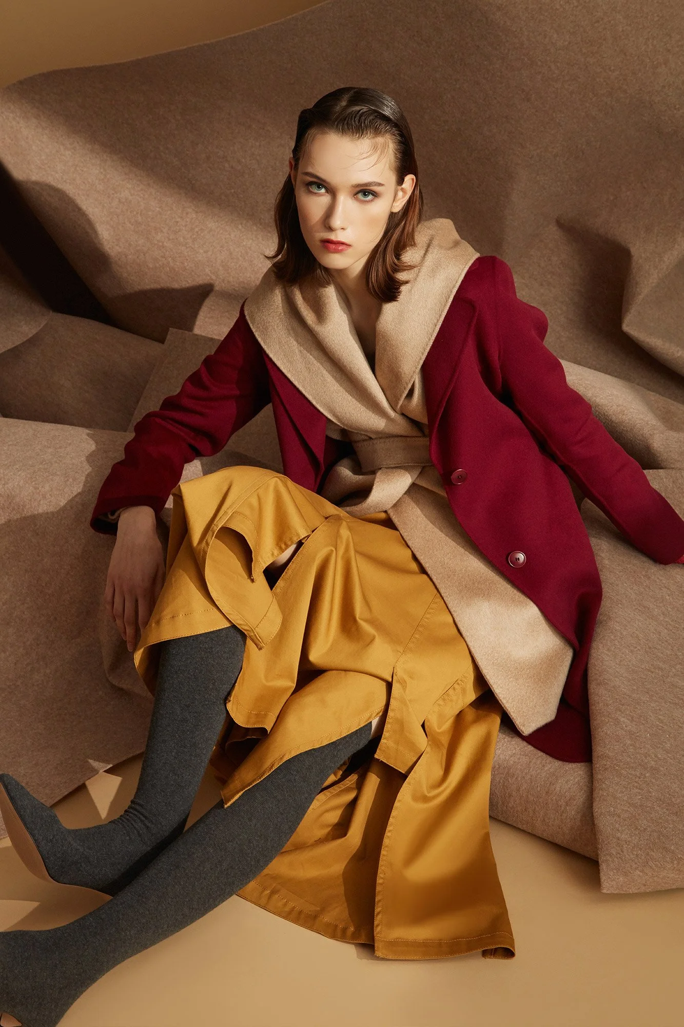 A woman sitting on a beige sofa, wearing a red coat with a beige hood, a yellow skirt, gray tights, and gray pointed shoes.