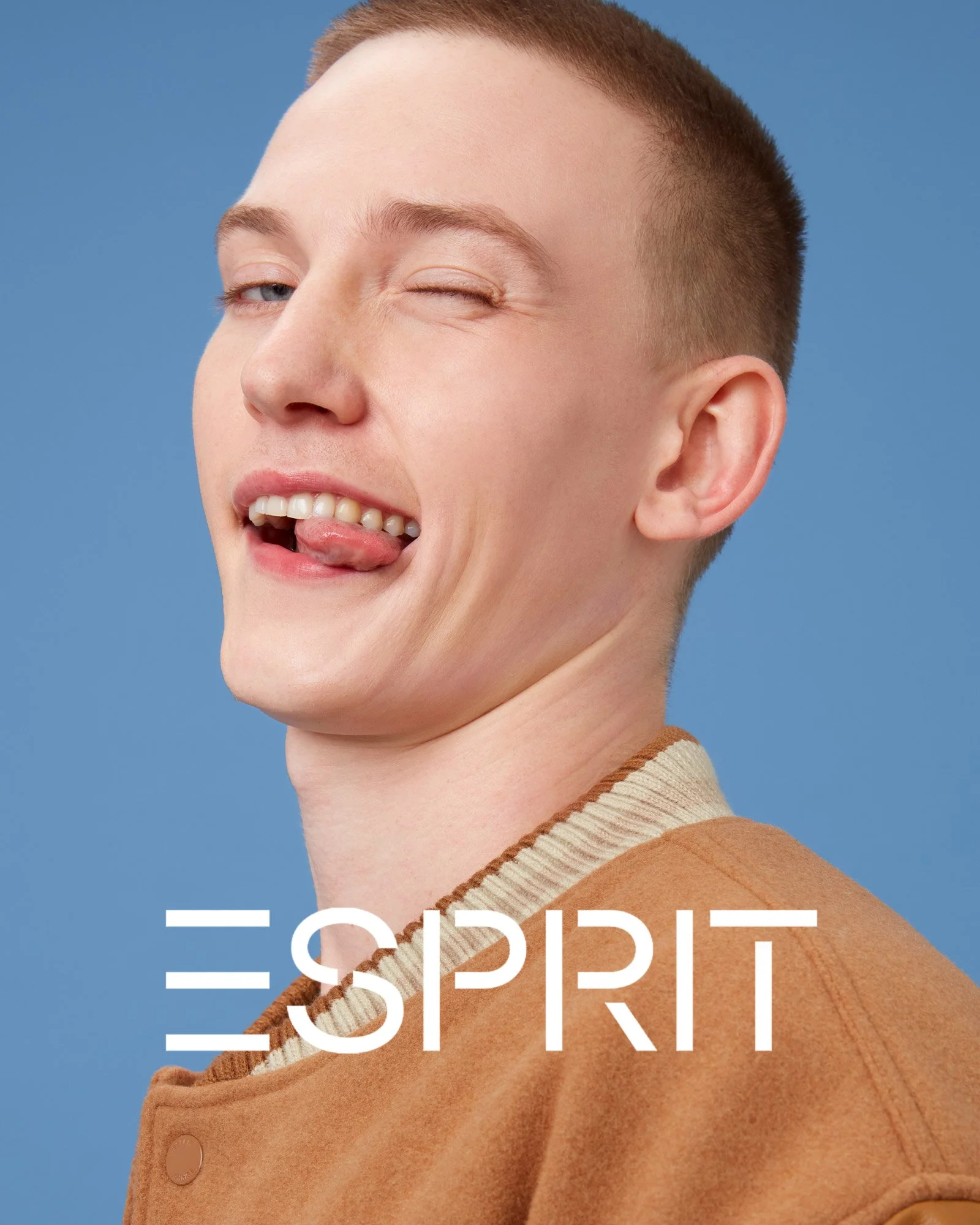 A young man with short hair winking and sticking out his tongue, wearing a tan jacket against a blue background, with the Esprit logo overlaid.