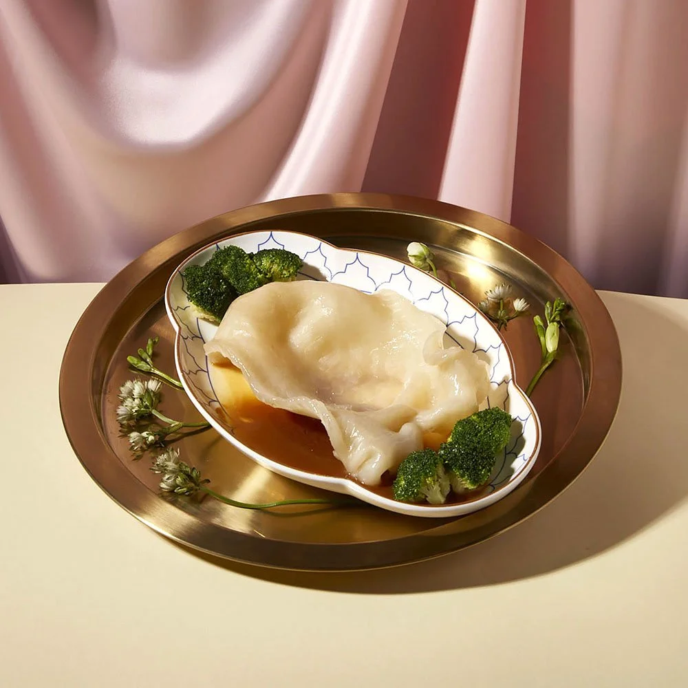 A plate with a steamed dumpling served with broccoli and garnished with small white flowers on a gold tray with a pink satin backdrop.