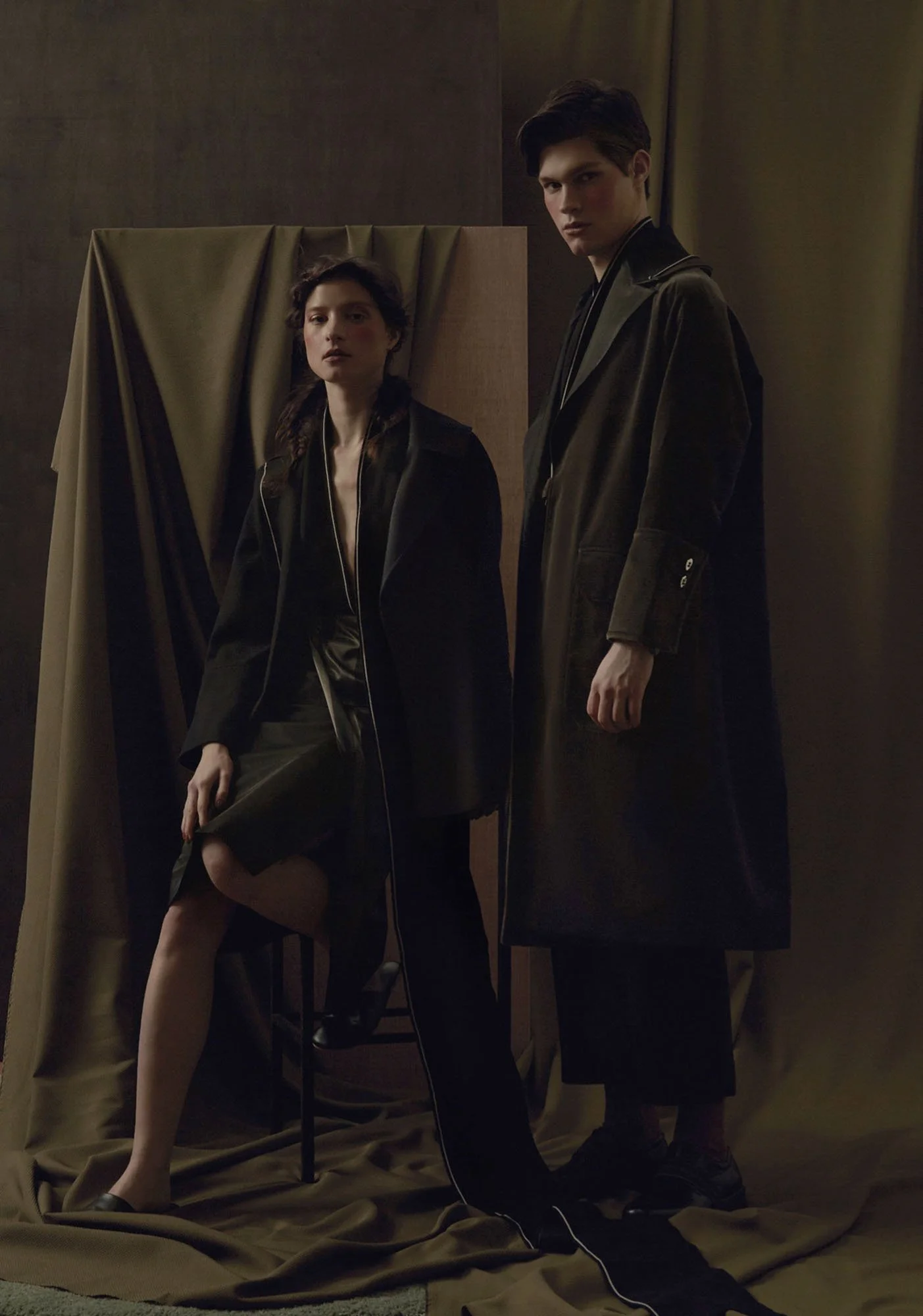 Two young models, a woman seated and a man standing, dressed in dark fashion coats, posed against a backdrop of brown and tan drapery with a dark background.