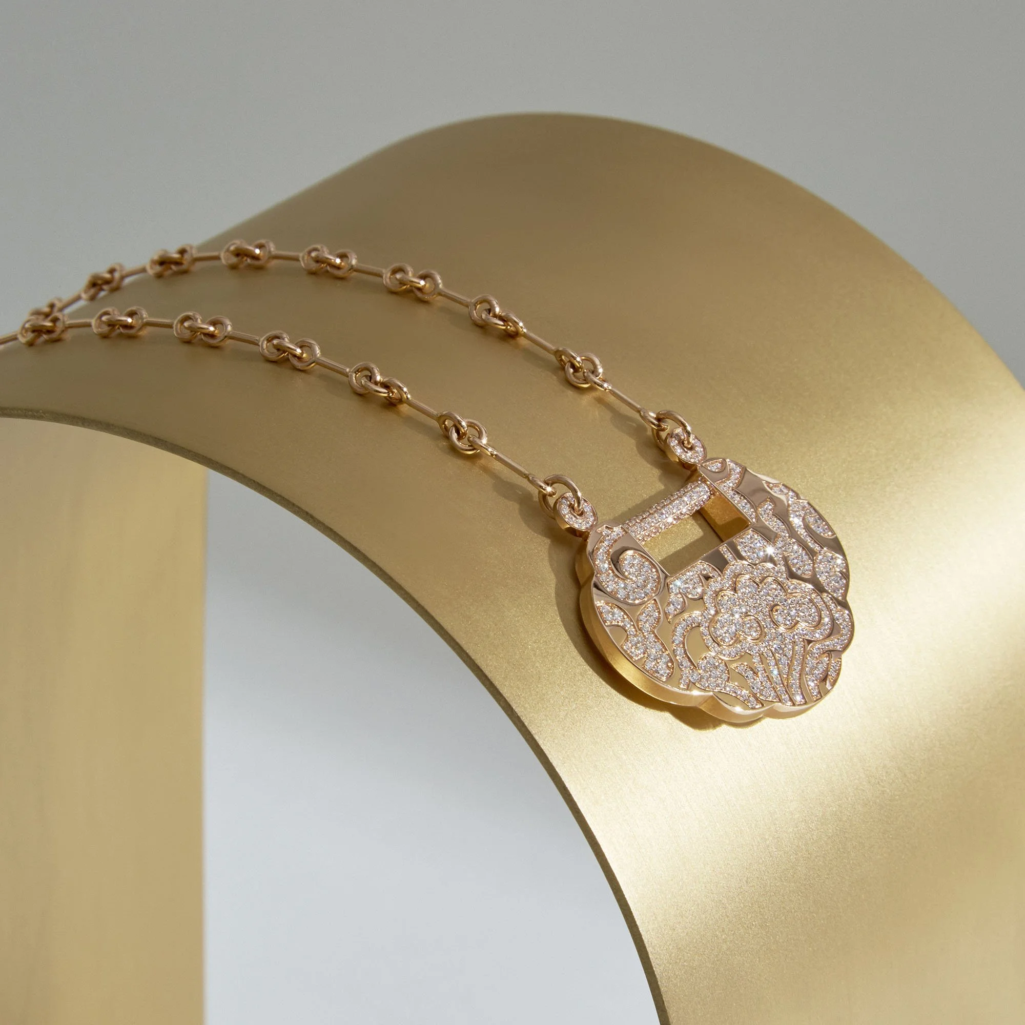 Gold necklace with ornate pendant encrusted with small diamonds, resting on a curved gold surface.