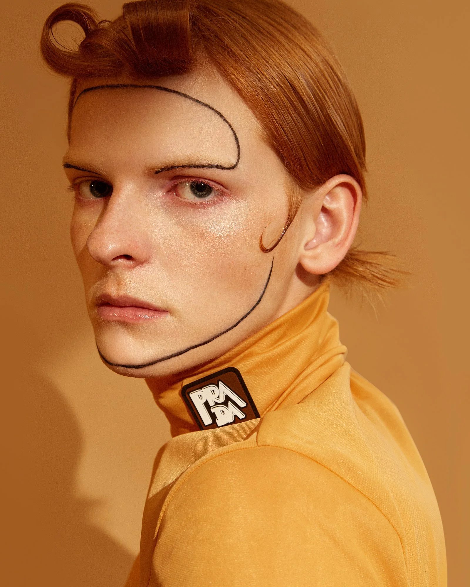 A person with short, red hair styled with a curl on top, wearing a yellow turtleneck with a black and white patch that reads 'PRADA' on the collar. They have curved black lines drawn on their face as part of a fashion or makeup look, against a yellow