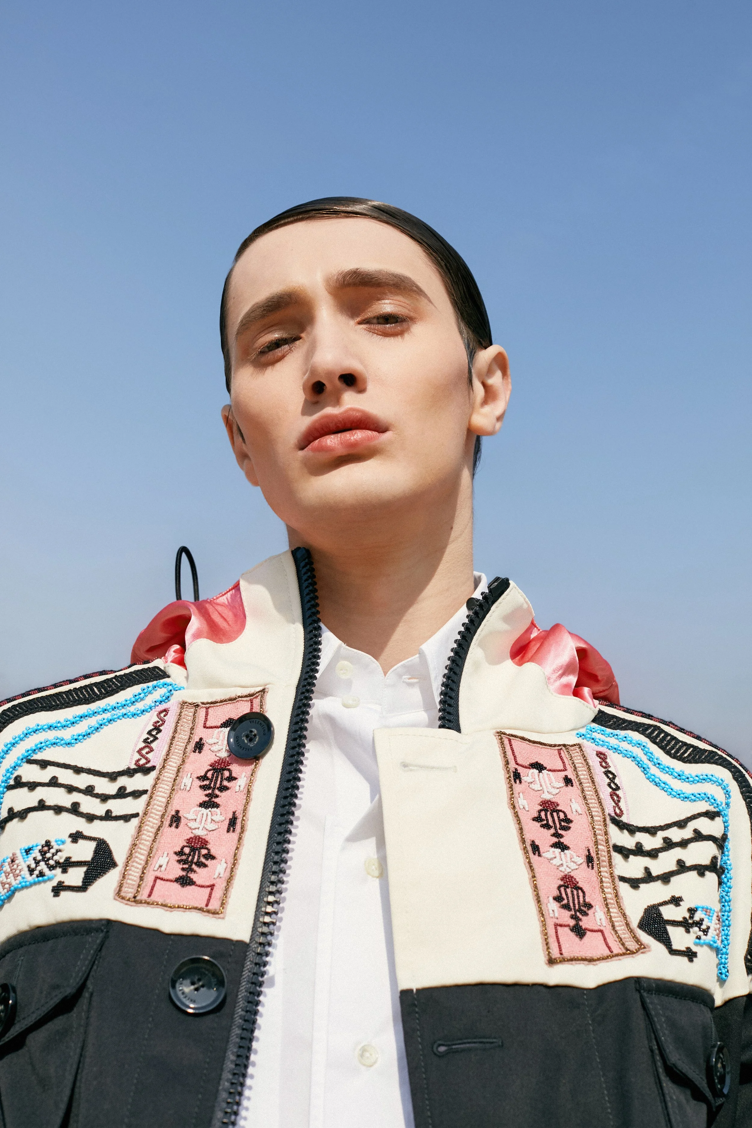 Close-up of a person with slicked-back hair wearing an embroidered jacket over a white shirt, standing against a clear blue sky.