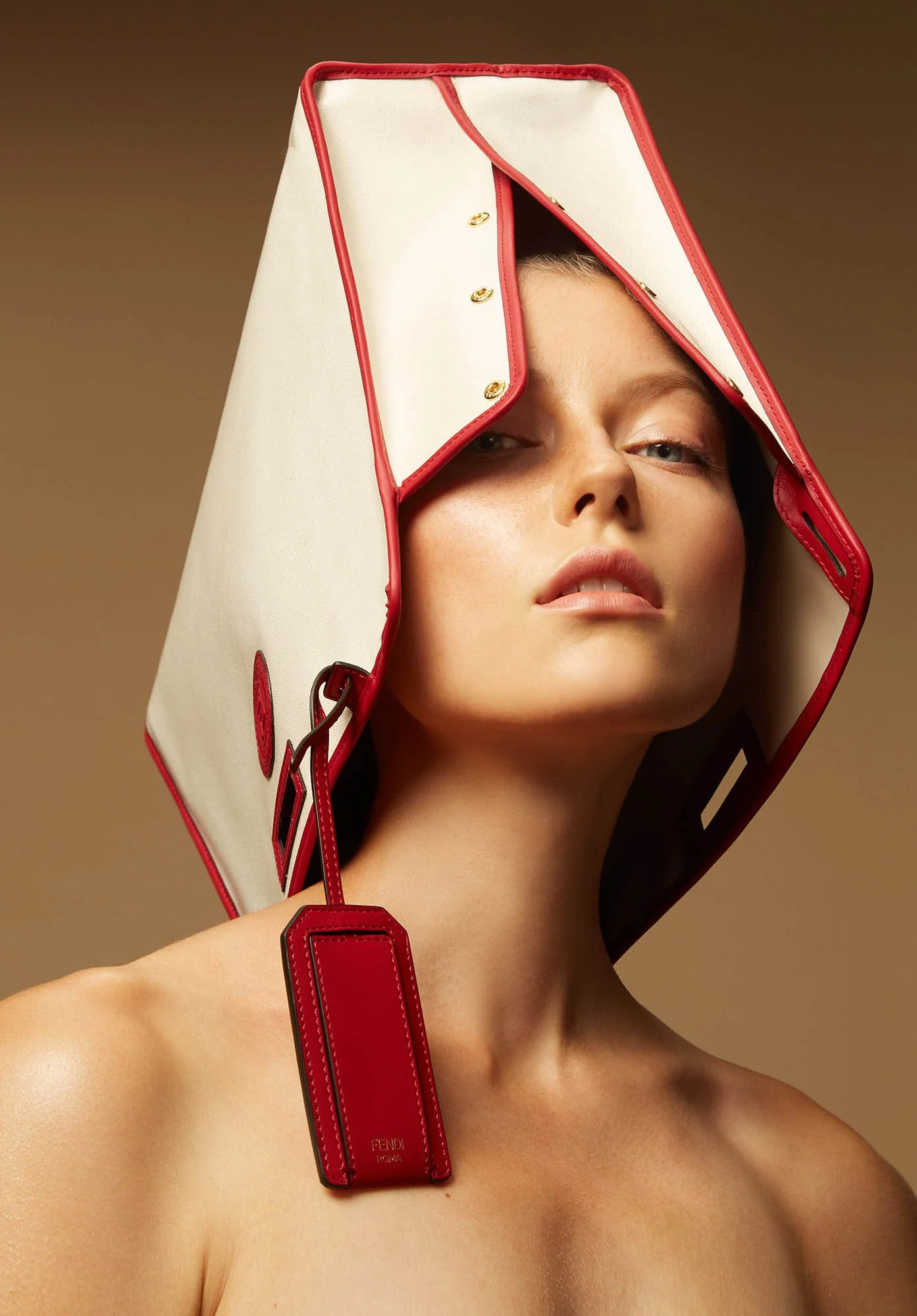 A woman with a beige complexion wears a large, boxy, white handbag with red trim and hardware on her head, partially covering her face against a neutral brown background.