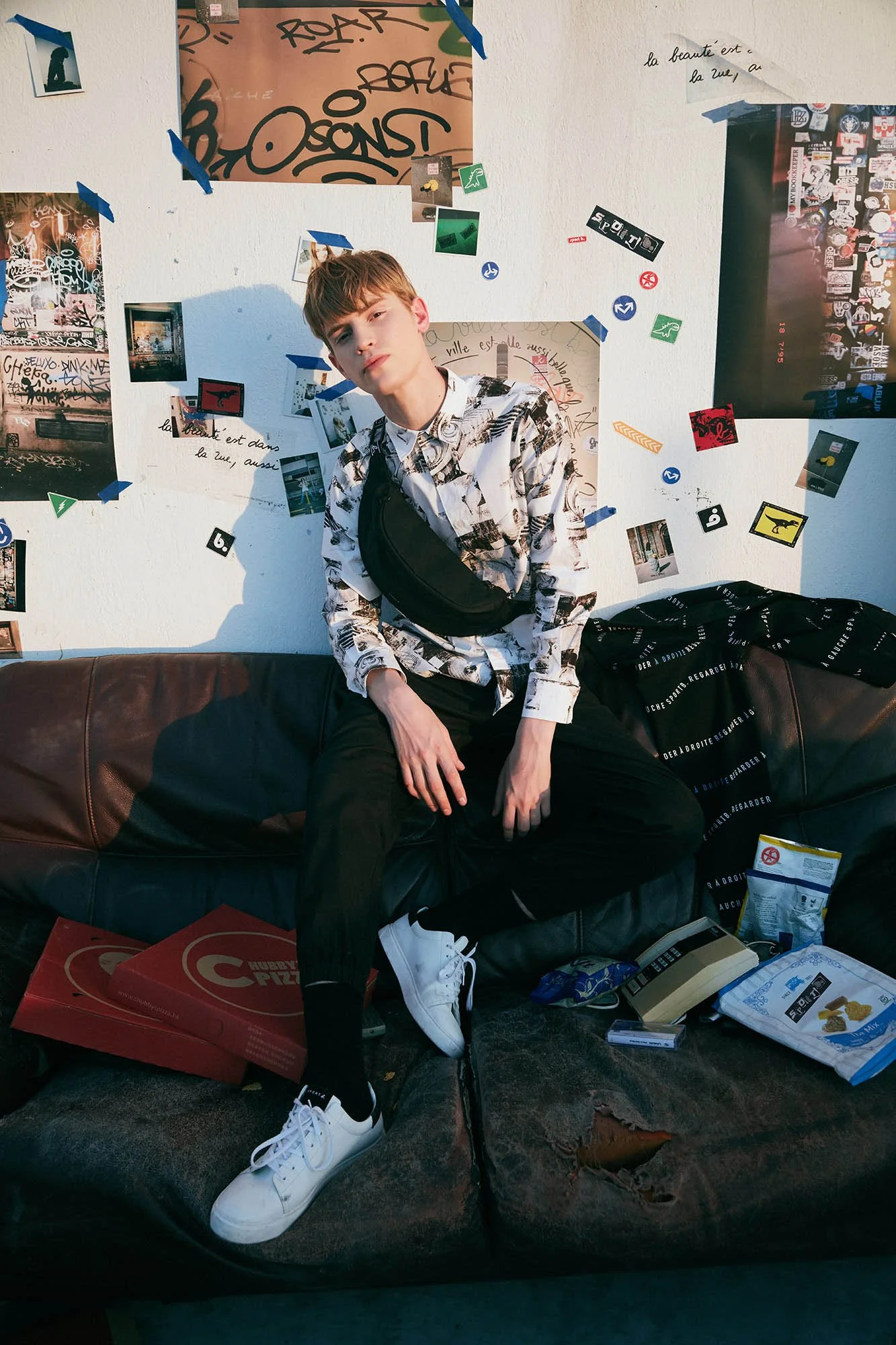 A young man with light skin and short, light brown hair sitting on a worn black leather couch in front of a white wall decorated with pictures, posters, and handwritten notes. He is wearing a black and white graphic shirt, black pants, white sneakers
