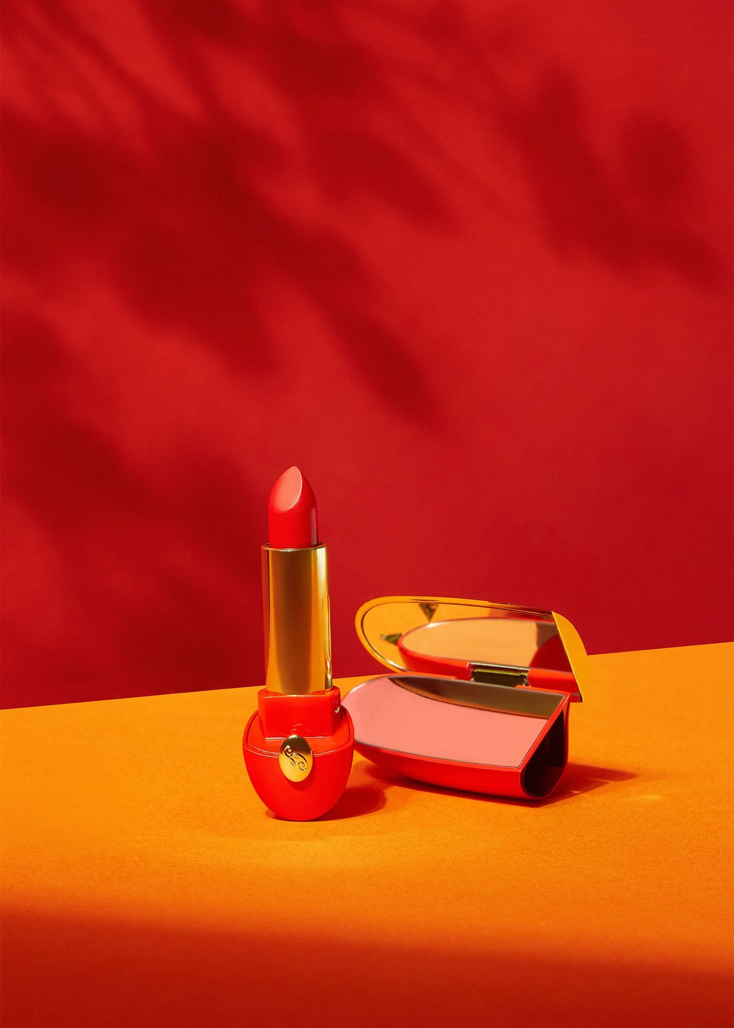A lipstick in a gold case with a pinkish red color standing upright next to a heart-shaped mirror compact, set against an orange surface and a red background.