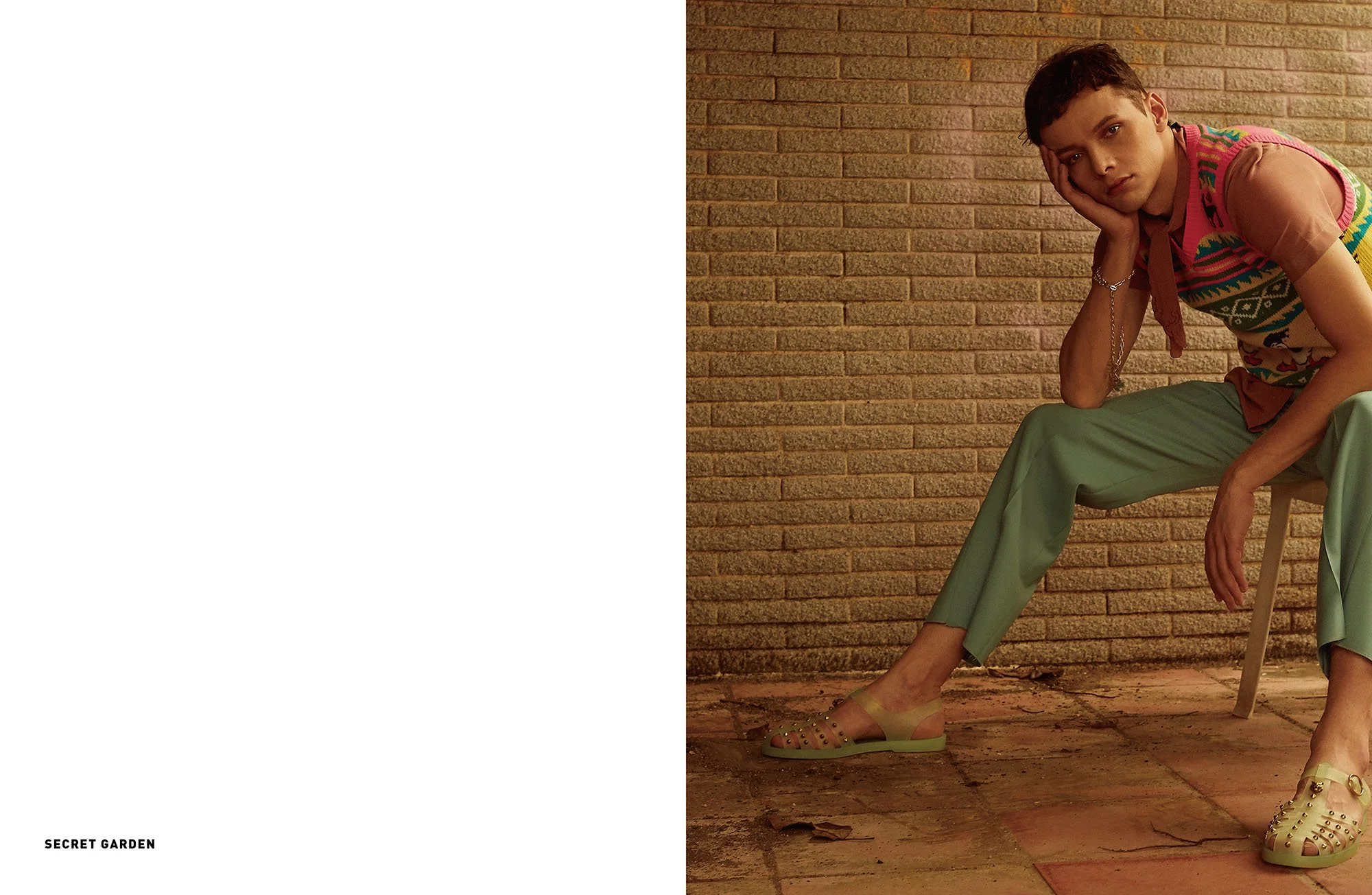 Young person sitting on a chair against a brick wall, wearing a colorful sweater vest, pink shirt, light green pants, and beige open-toe shoes, with a contemplative expression and resting their head on their hand.